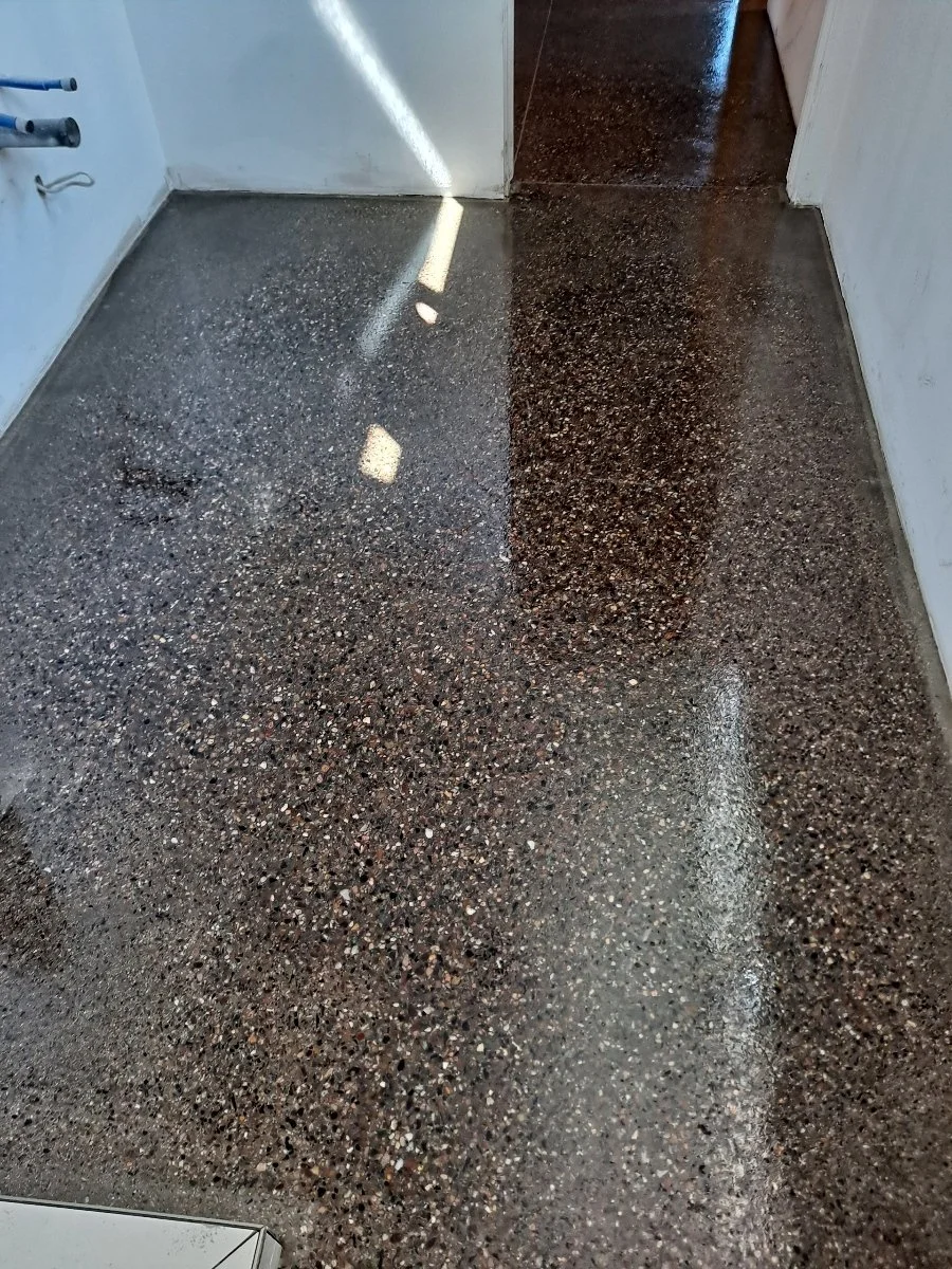 A freshly polished, speckled concrete floor in a room with white walls and a door, showing a bright reflection of light.