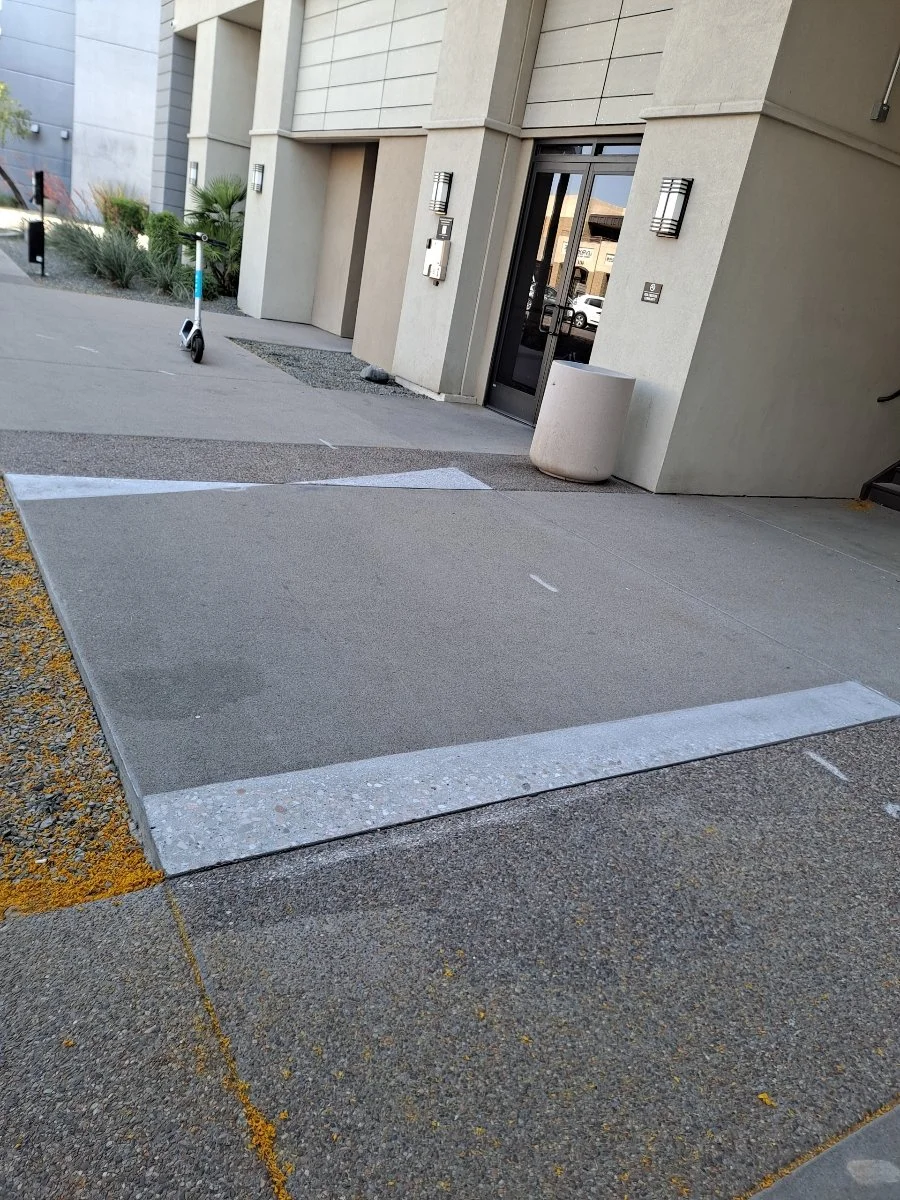 Sidewalk ramp with textured surface for wheelchair access outside a modern building, with a scooter parked nearby and some plants in the background.