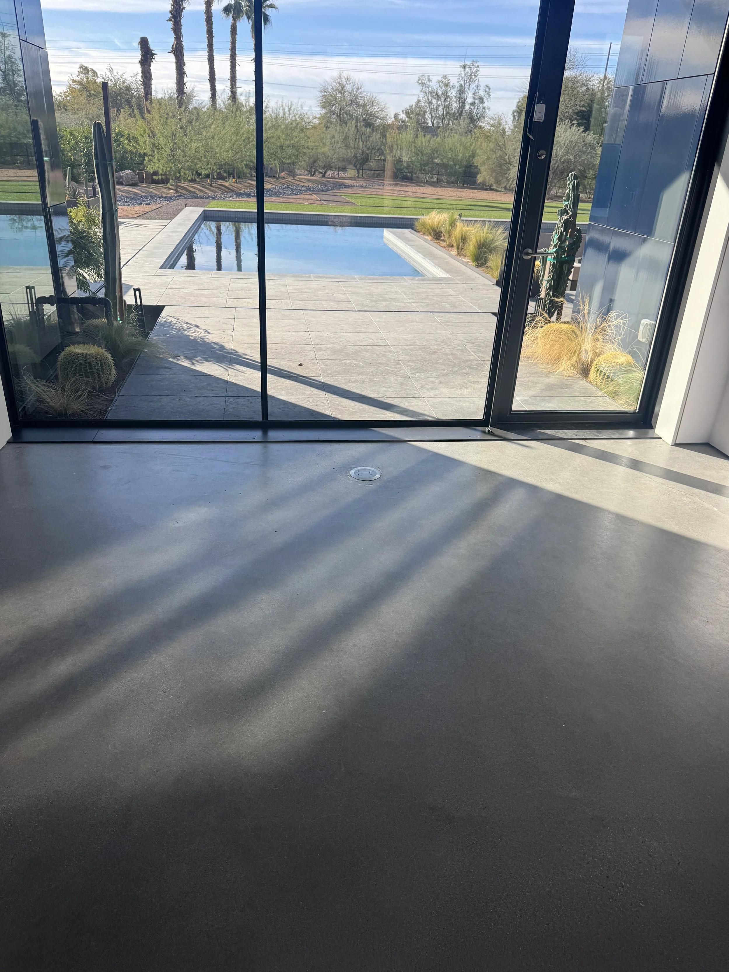 View through glass doors showing a backyard with a rectangular swimming pool, green grass, trees, and a clear blue sky.