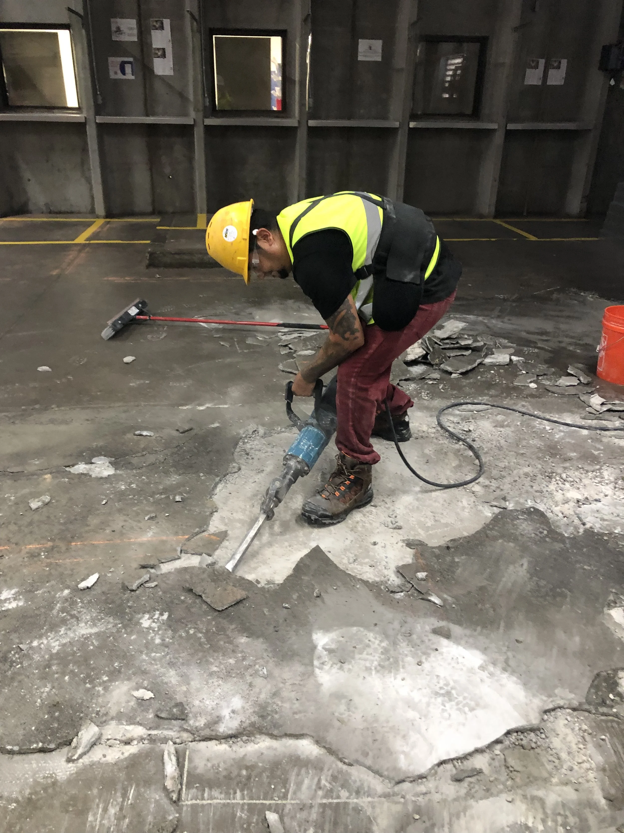 A construction worker wearing a yellow safety helmet, a reflective safety vest, a black shirt, red pants, and brown boots uses a jackhammer to break up concrete on a construction site. There is an orange bucket and a squeegee in the background.