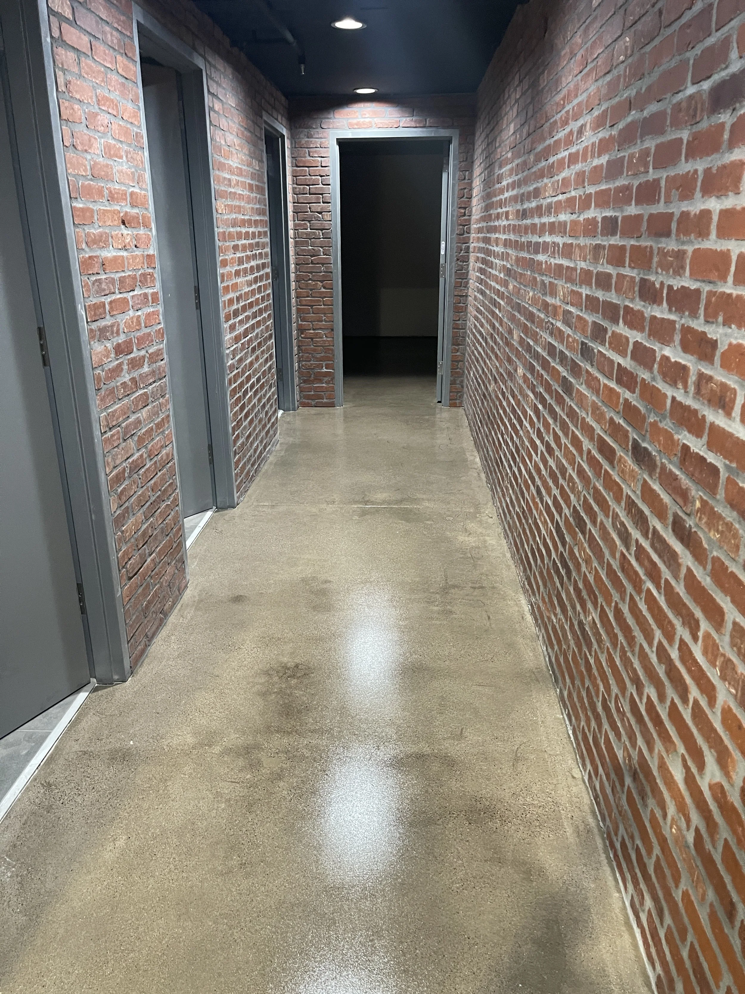 A hallway with red brick walls, a polished concrete floor, and a black ceiling with built-in lighting. There are doors on the left side and an open doorway at the end.