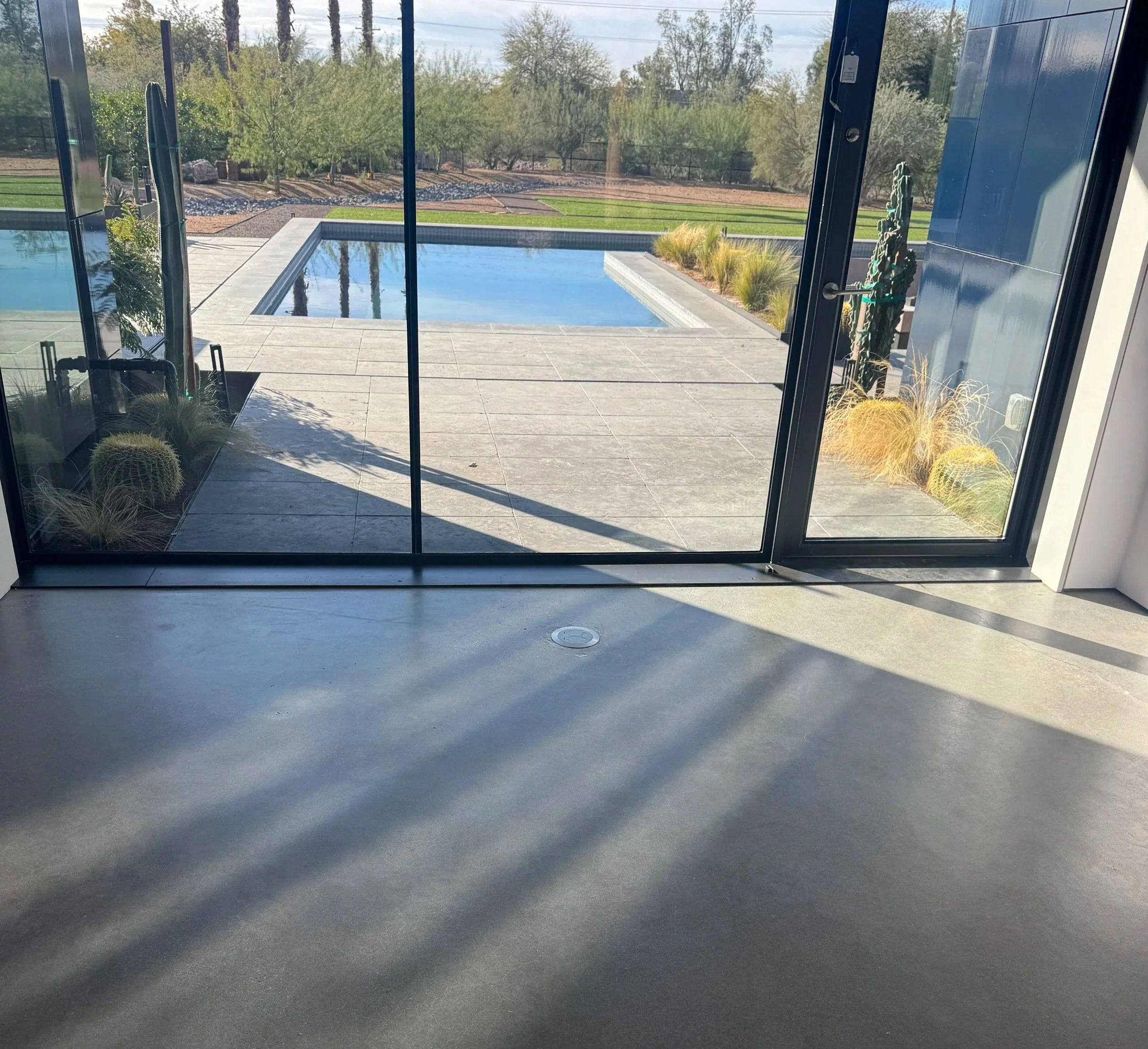View of an outdoor swimming pool through glass sliding doors, with a concrete patio, desert plants, and a grassy area with trees in the background.