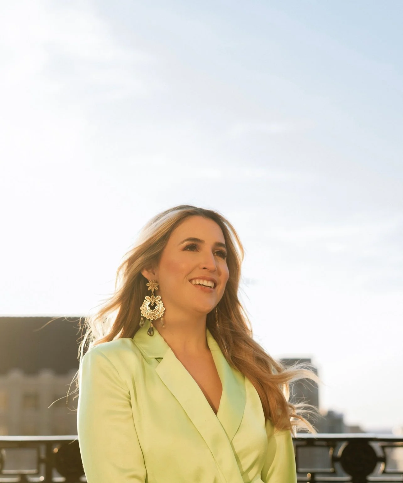 A woman with long hair wearing a light yellow blazer and large decorative earrings, smiling outdoors during sunset, with city buildings in the background.