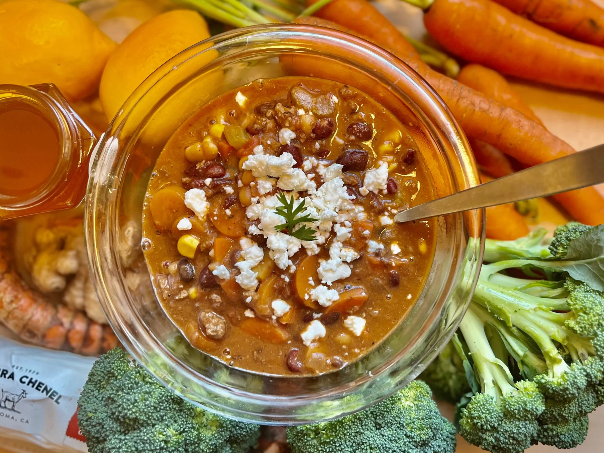 A bowl of chili topped with cheese and cilantro, surrounded by fresh vegetables including broccoli, carrots, yellow potatoes, and a bunch of carrots.