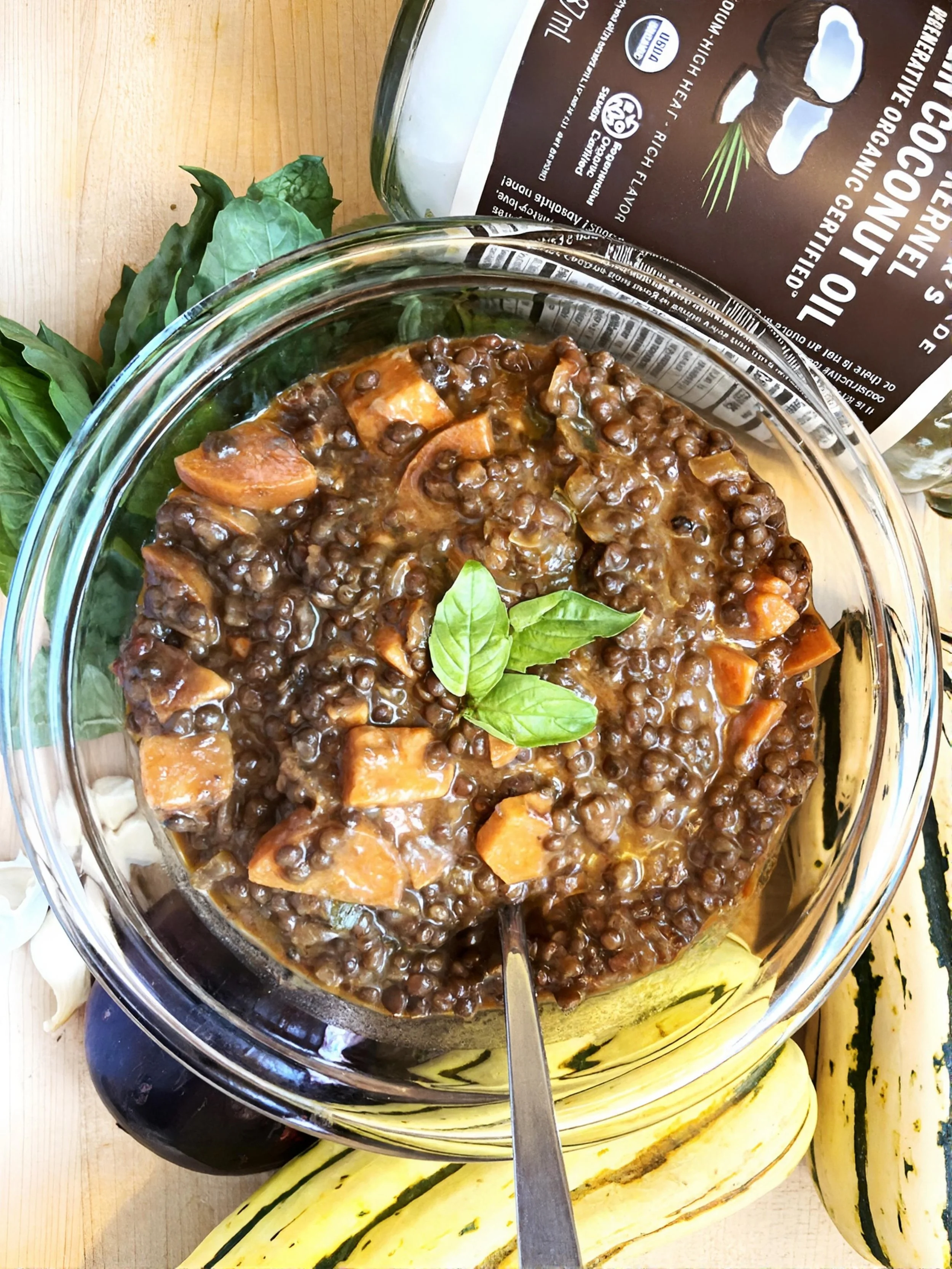 A bowl of lentil stew with carrots and basil leaves on top, with sweet potatos and a package of organic mushrooms in the background.