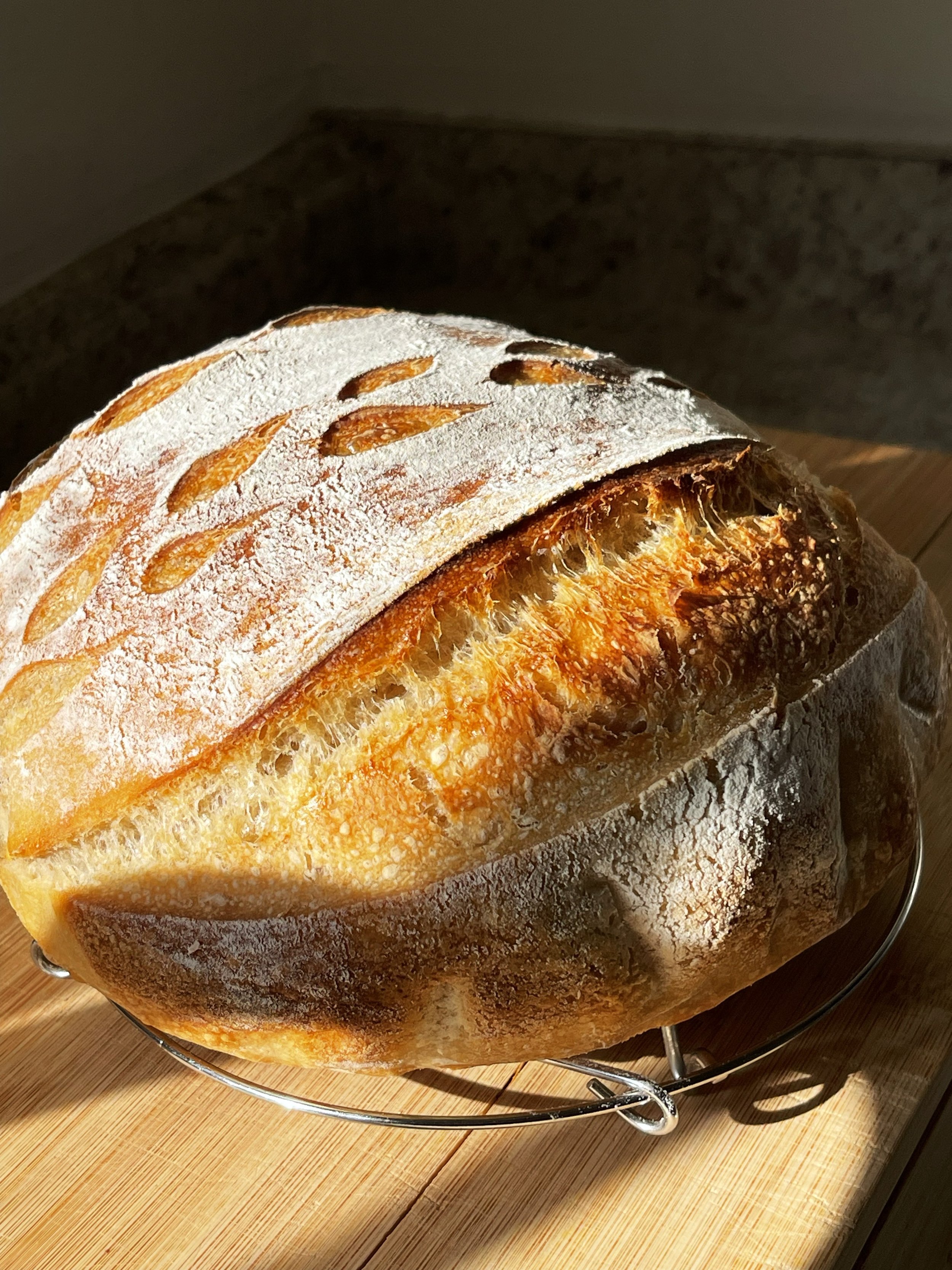 A loaf of crusty bread with a dusting of flour on top, resting on a wire rack on a wooden surface.