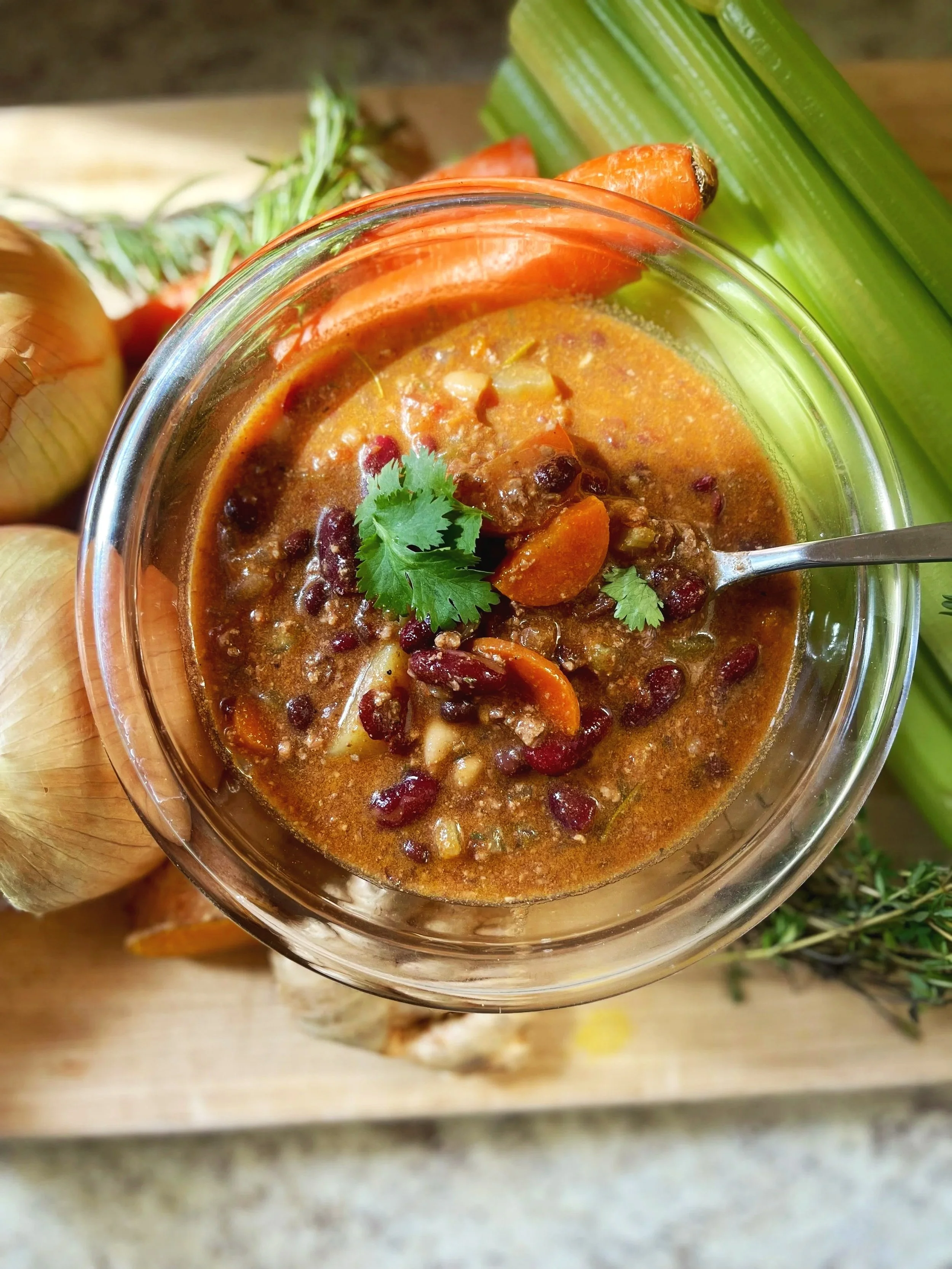 A bowl of homemade chili with beans, chunks of vegetables, and herbs, placed on a wooden surface with celery and onions nearby.