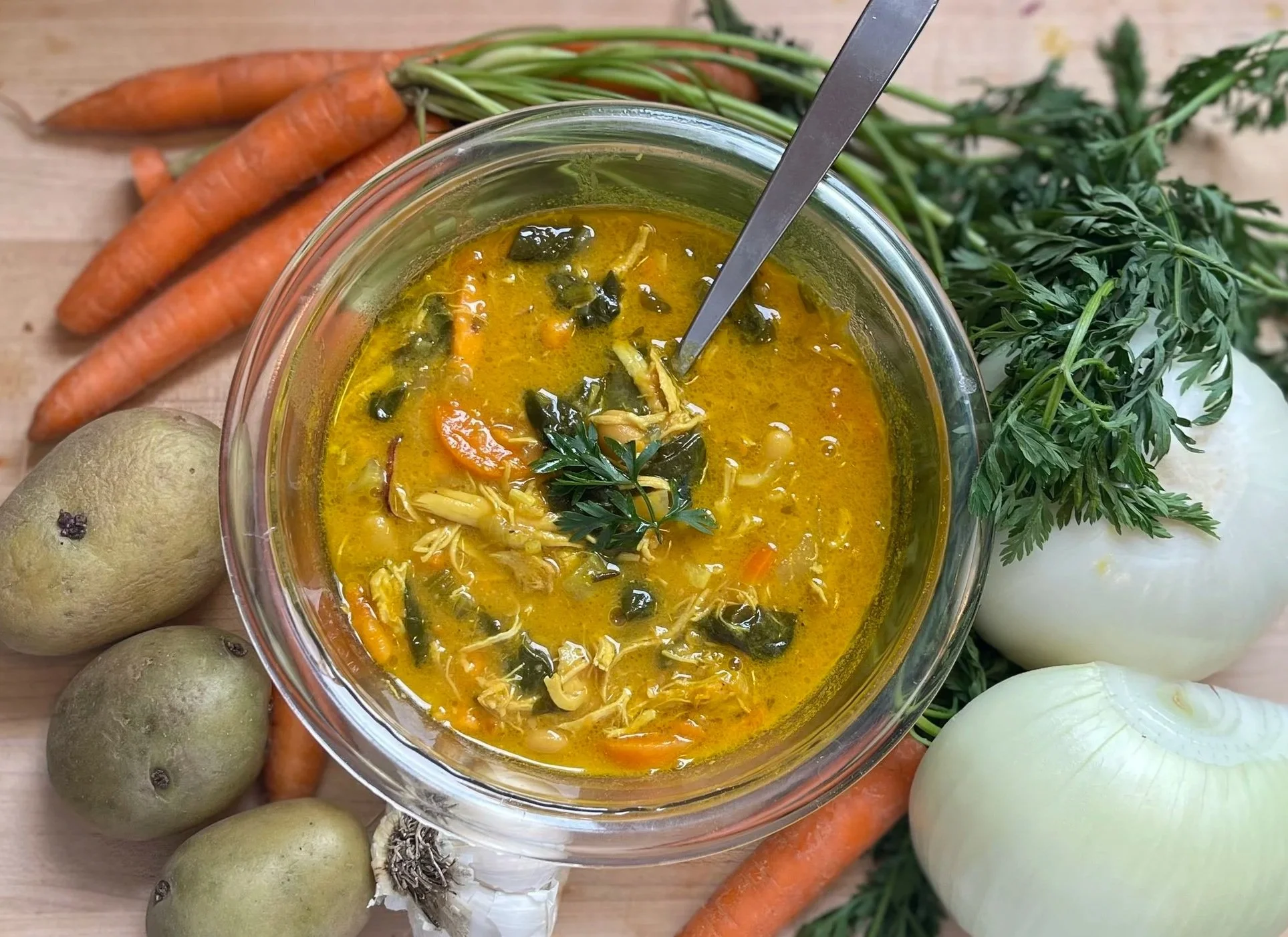A bowl of homemade vegetable soup with carrots, zucchini, and leafy greens, garnished with parsley, surrounded by fresh carrots, onions, and potatoes on a wooden surface.