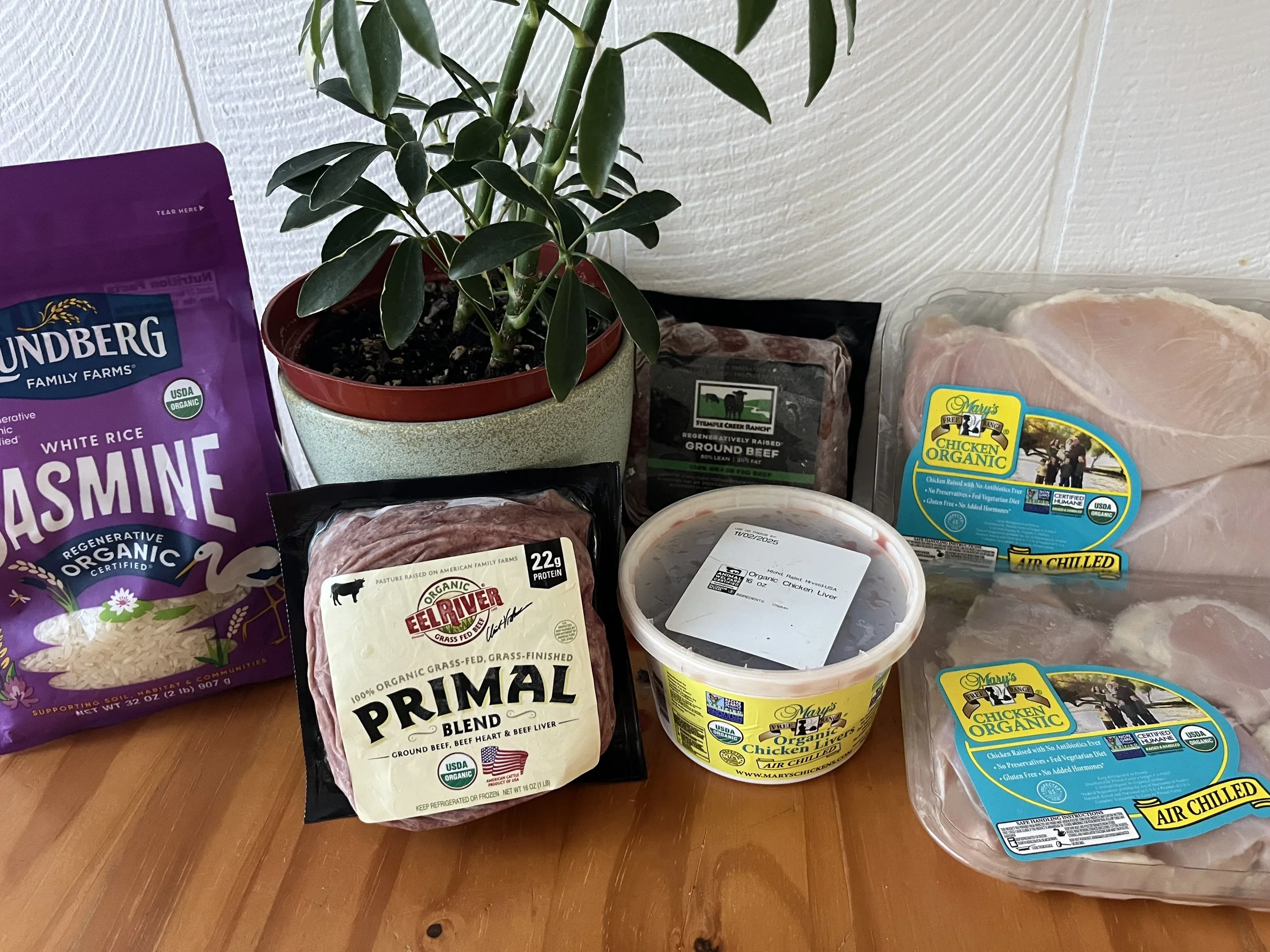 Assorted fresh meats including ground beef, chicken liver, and chicken pieces, a bag of jasmine rice, and a potted green plant on a wooden surface.