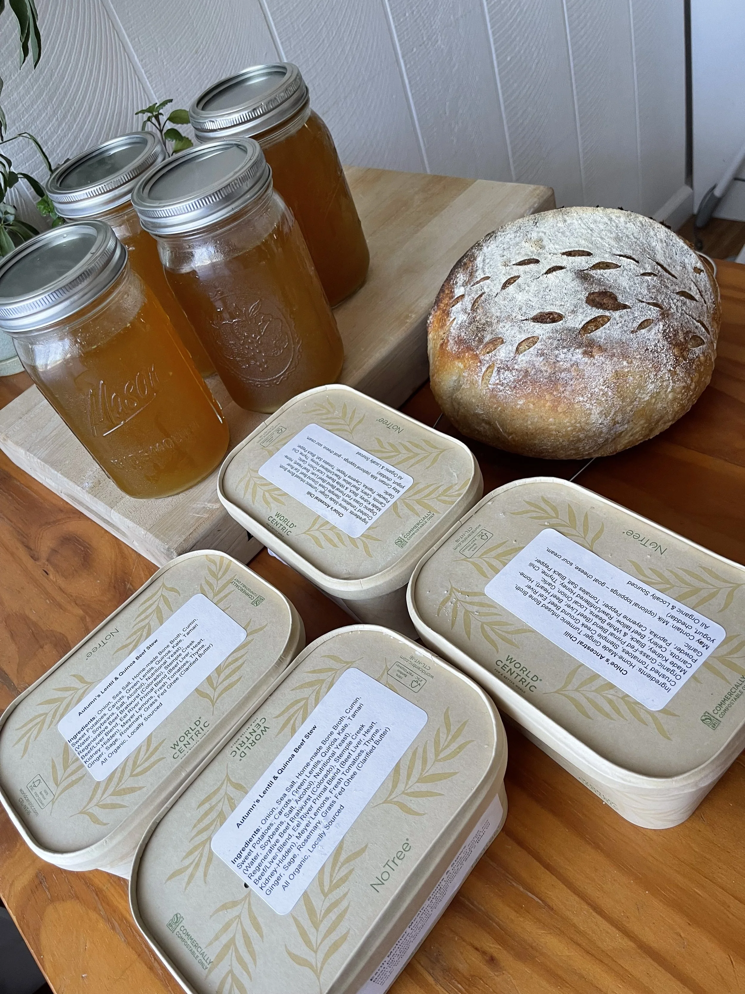Four jars of bone broth, a round loaf of homemade sourdough bread, and four containers of Earth Mother Meals Soups and Stews on a wooden table.