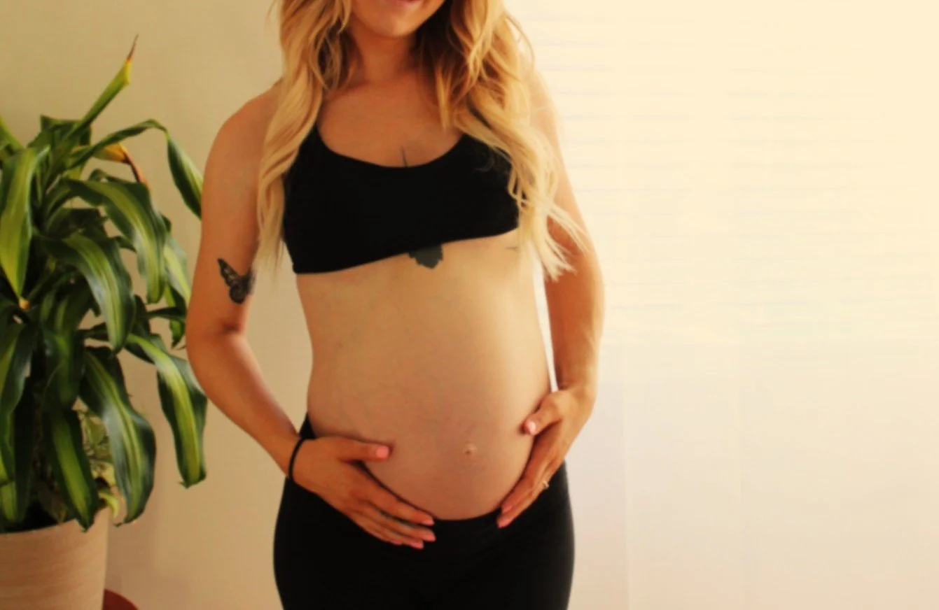 A pregnant woman with long blonde hair, wearing a black sports bra and black pants, standing near a potted plant in a well-lit room.