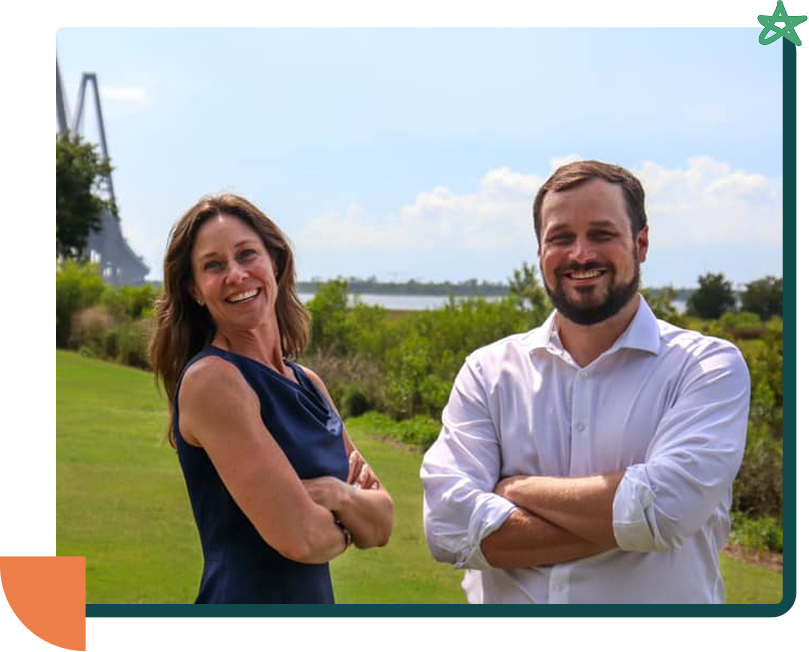 A smiling woman in a sleeveless navy dress and a smiling man in a white shirt with rolled-up sleeves standing outdoors near a body of water and greenery, with a bridge in the background.