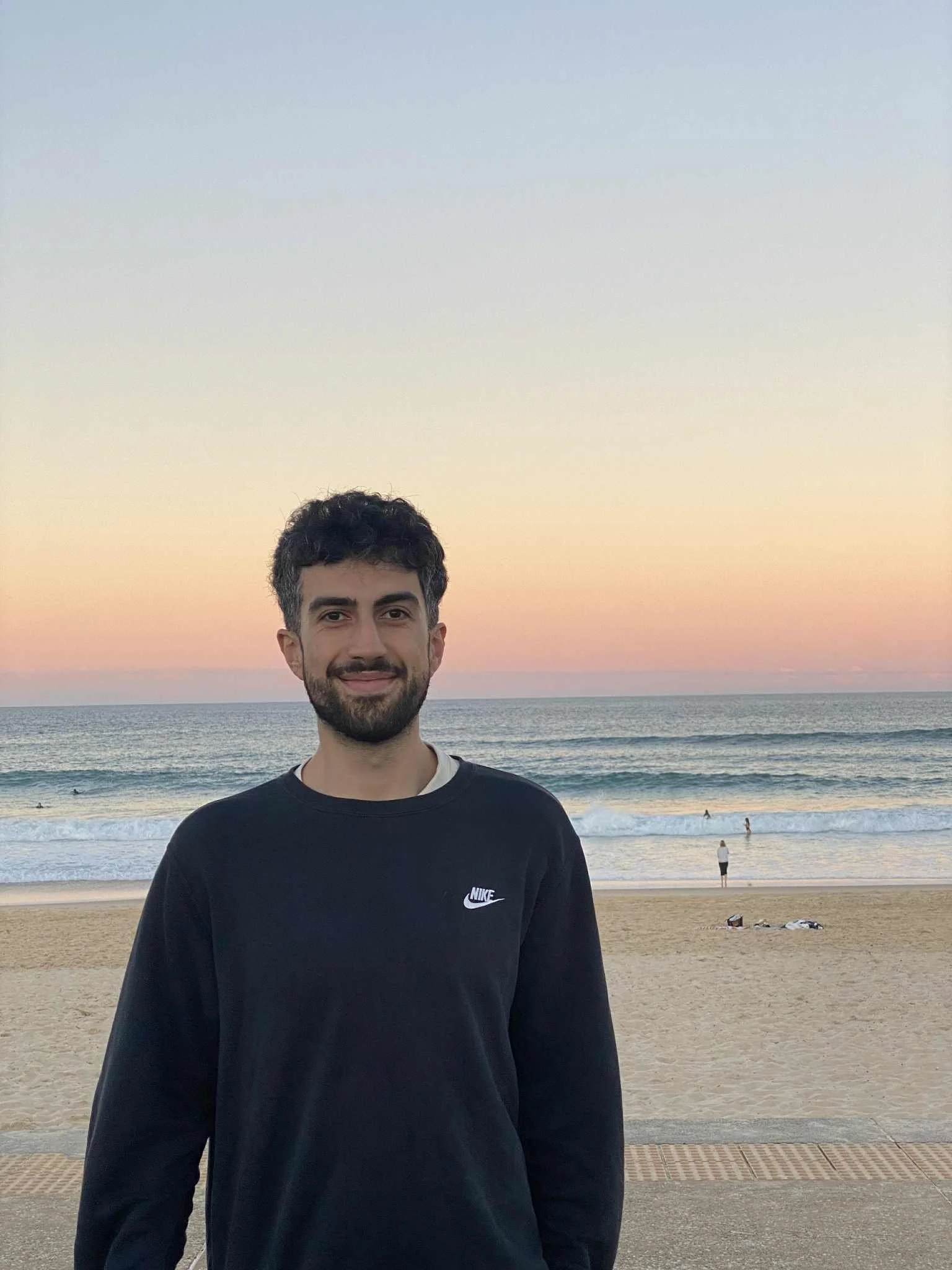 A young man with dark curly hair and a beard smiling at the camera, standing on a beach at sunset with the ocean and a few people in the background.