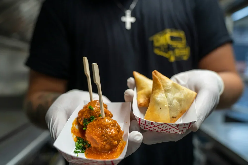 Person wearing black shirt and white gloves holding two paper trays of Indian food; one with two samosas and the other with meatballs in tomato sauce garnished with cilantro.