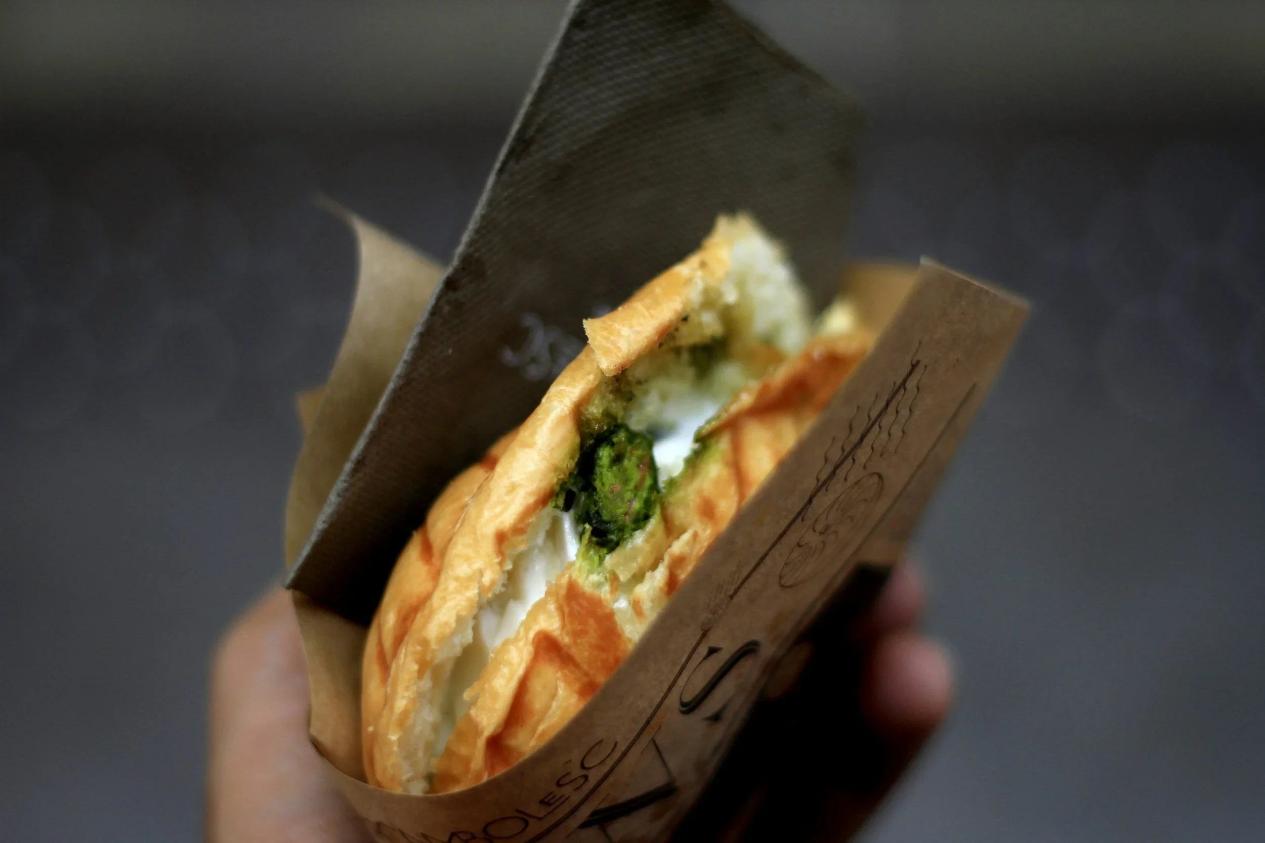 A hand holding a sandwich with fried bread, green spinach, and white cheese inside, wrapped in brown paper.