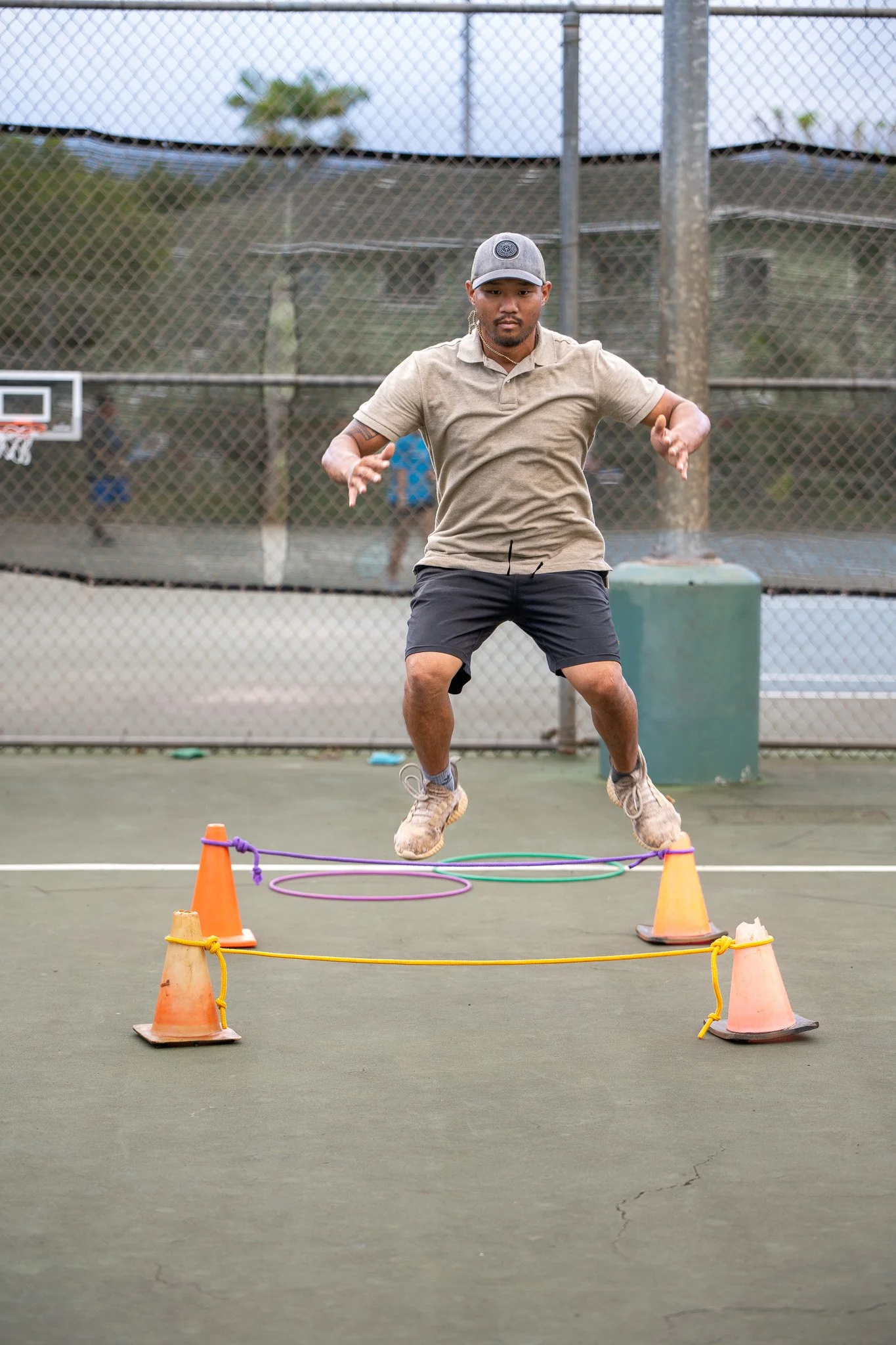 Man jumping over hurdles with cones and hoops on an outdoor tennis court.
