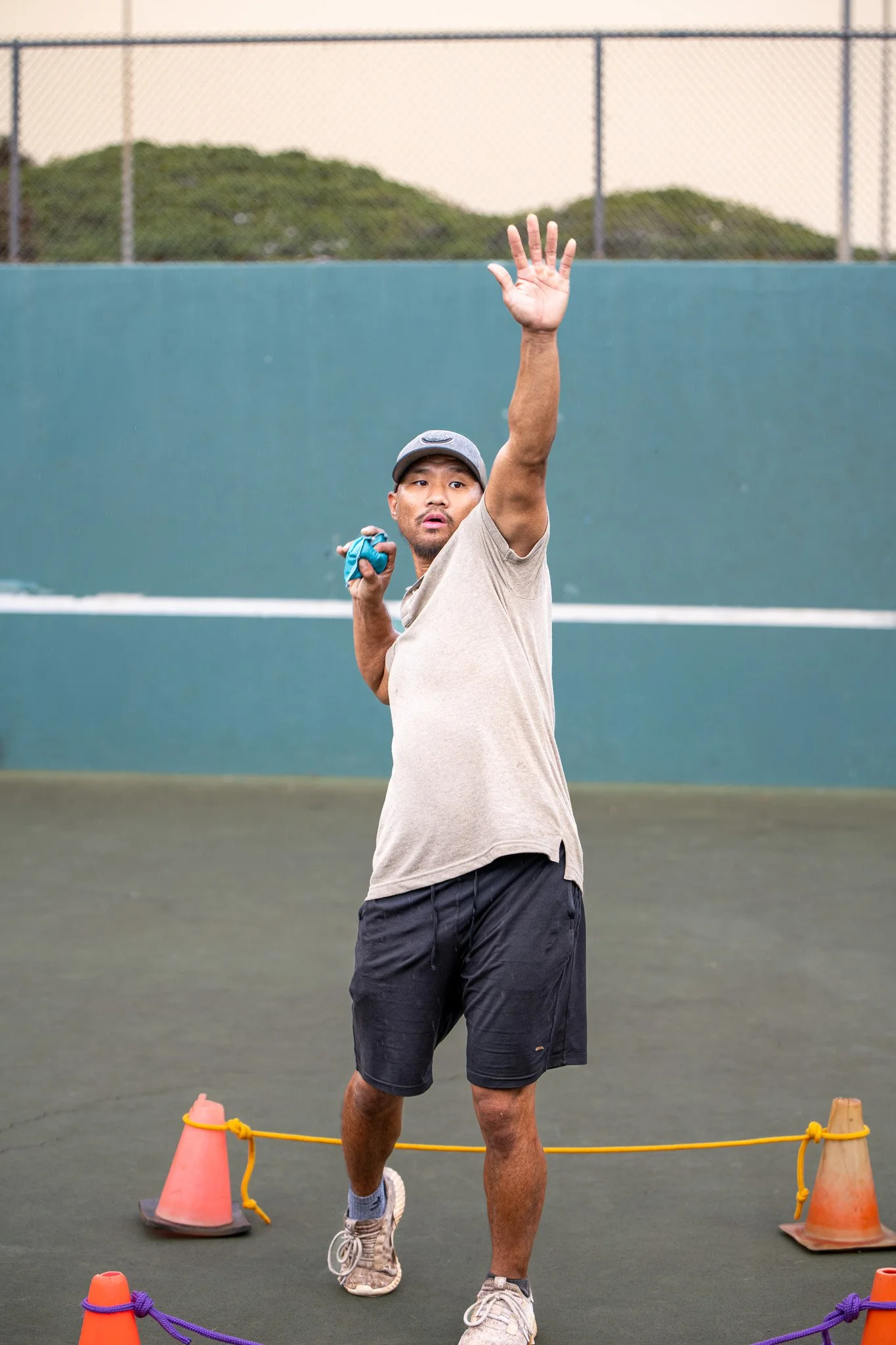 A man is preparing to serve or hit a tennis ball on a tennis court, wearing a gray cap, beige t-shirt, black shorts, and sneakers, holding a tennis ball in his left hand and a tennis racquet in his right hand, with orange cones and yellow ropes on the court.