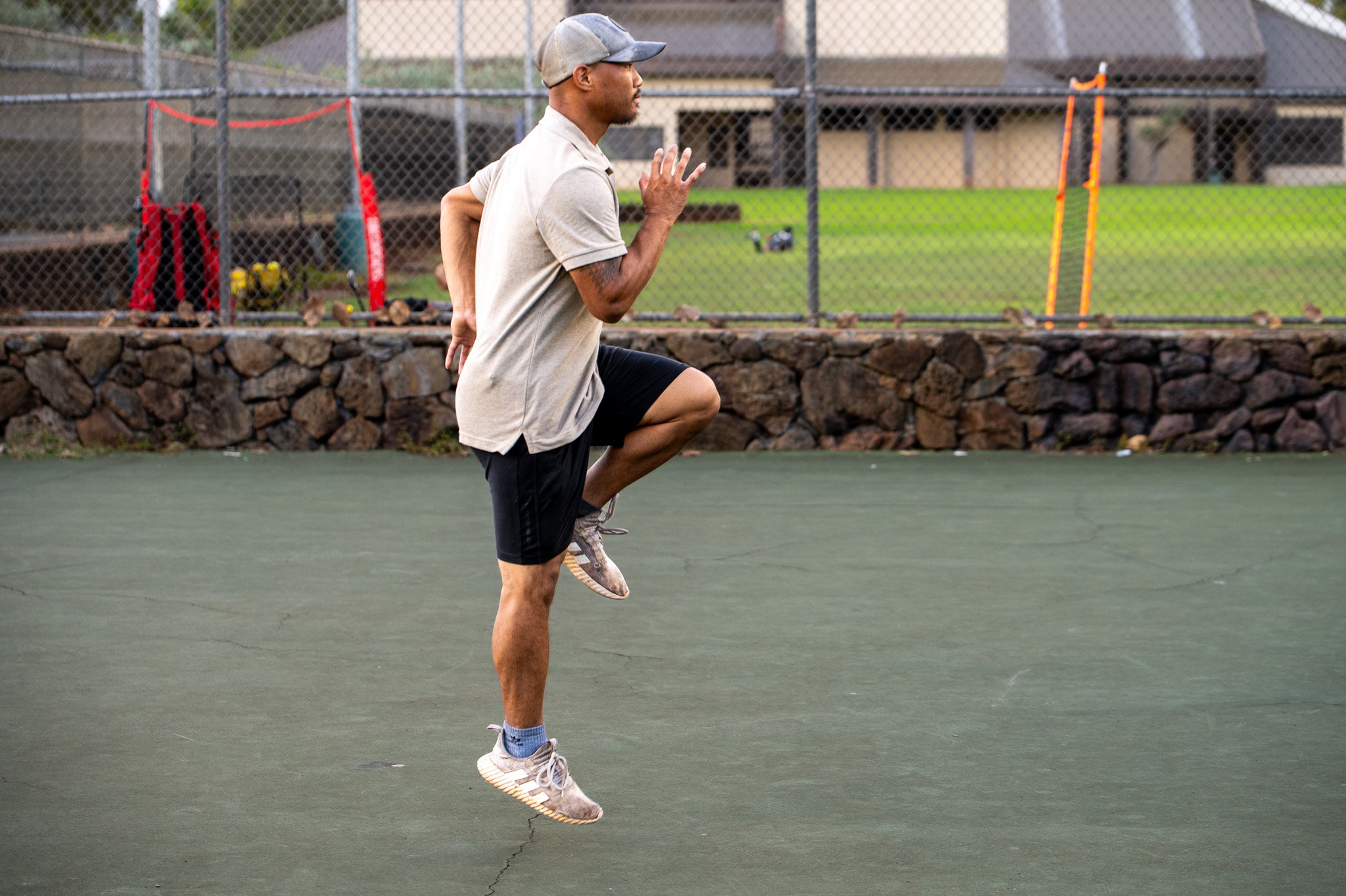 A man dressed in athletic clothing and a baseball cap is practicing a high knee exercise on an outdoor tennis court with a stone wall and a chain-link fence in the background.