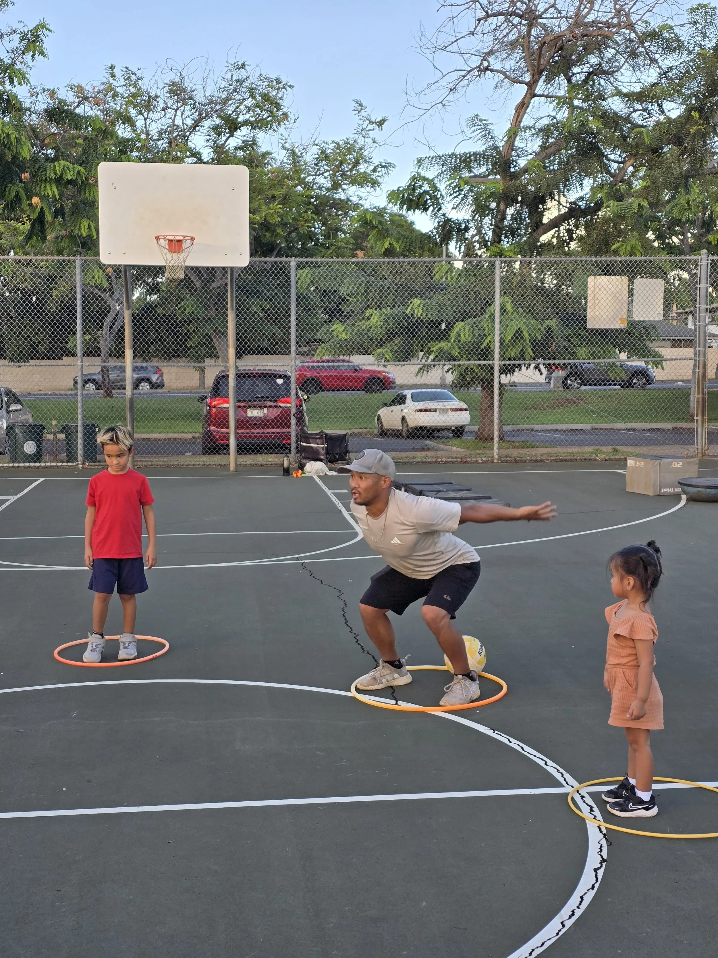 A man coaching two young children on a basketball court. The man is crouched with arms out, squatting in a yellow hoop, while the children stand in hula hoops, ready for activity. There is a basketball, parked cars, and trees in the background.