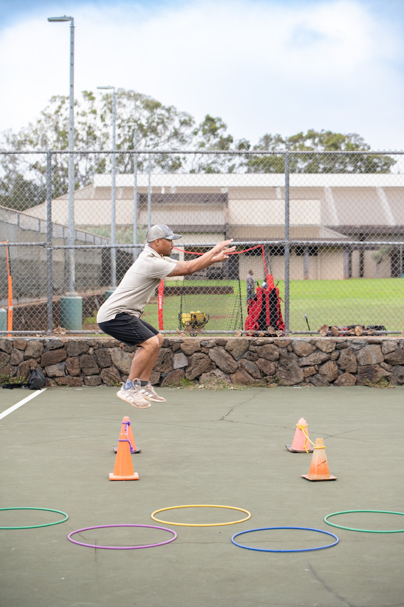 A man in athletic clothing jumping over a small obstacle course on a green outdoor court with hula hoops and traffic cones, with a chain-link fence and trees in the background.