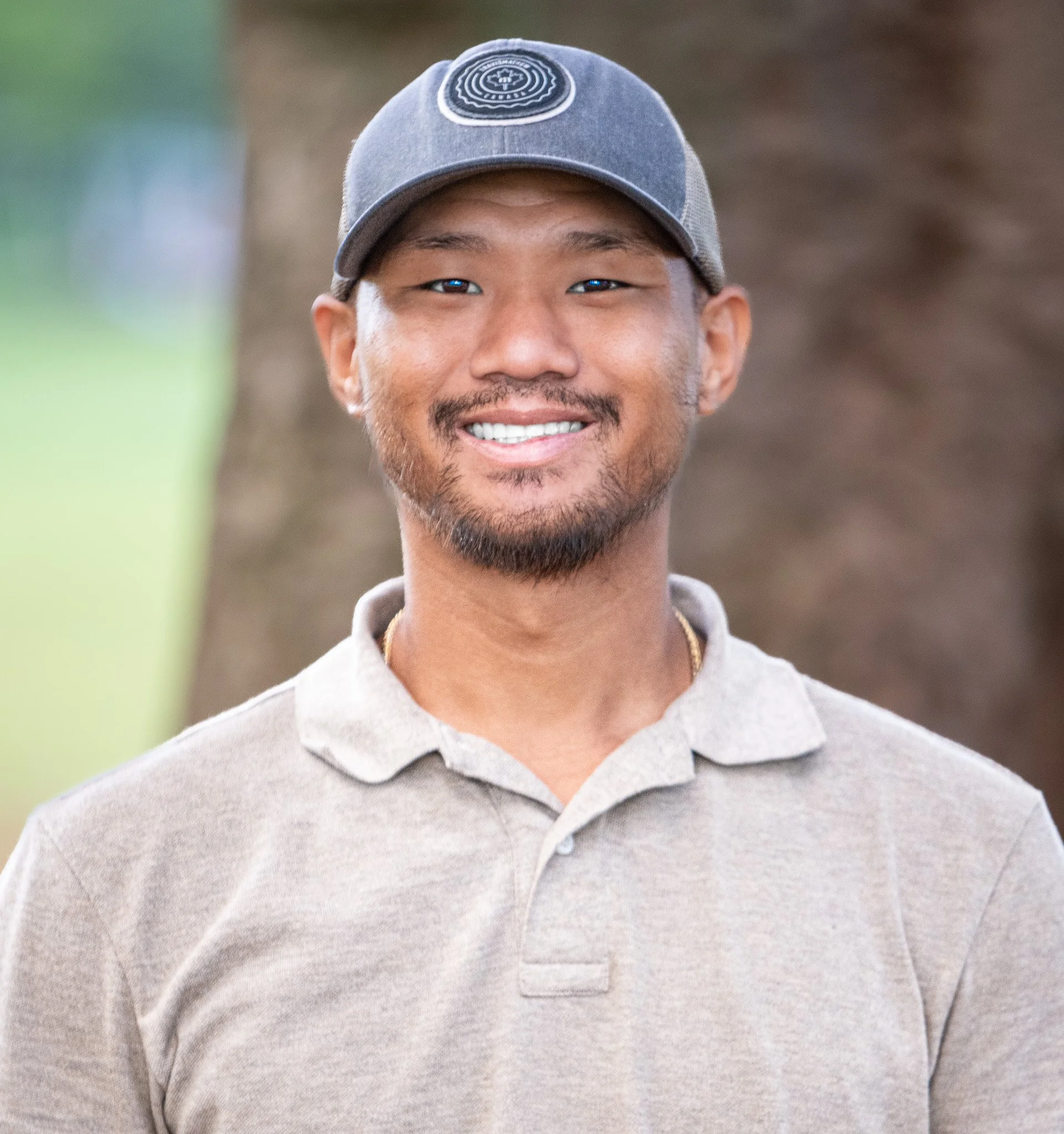 A smiling man wearing a gray baseball cap and beige polo shirt outdoors with blurred trees in the background.
