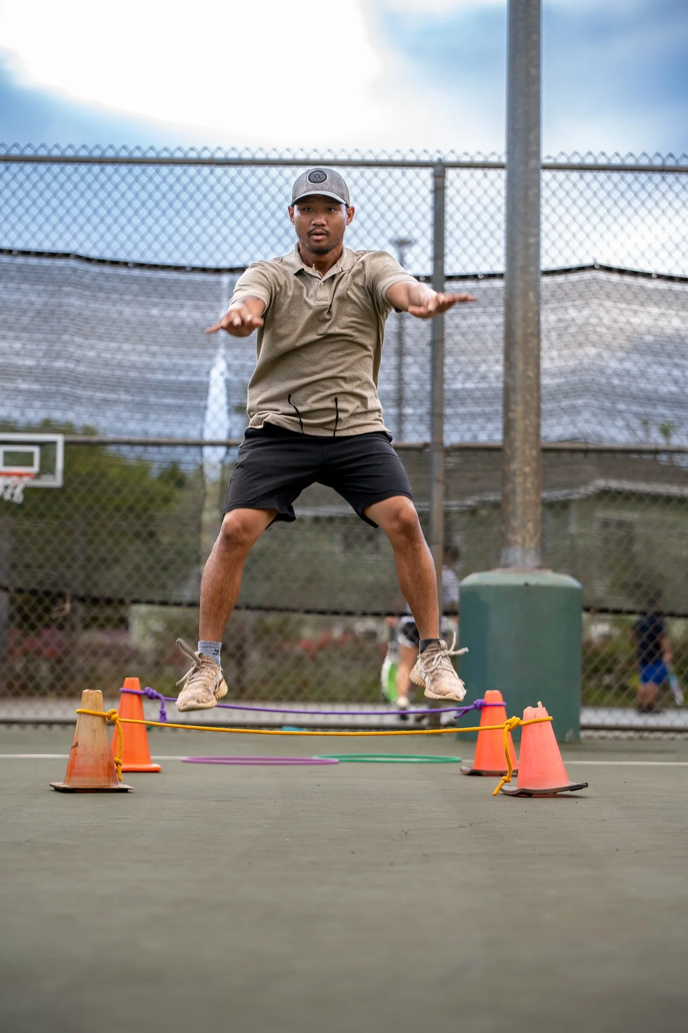 Young man jumping over a low obstacle on a tennis court during daytime, with cones and ropes marking the obstacle course.