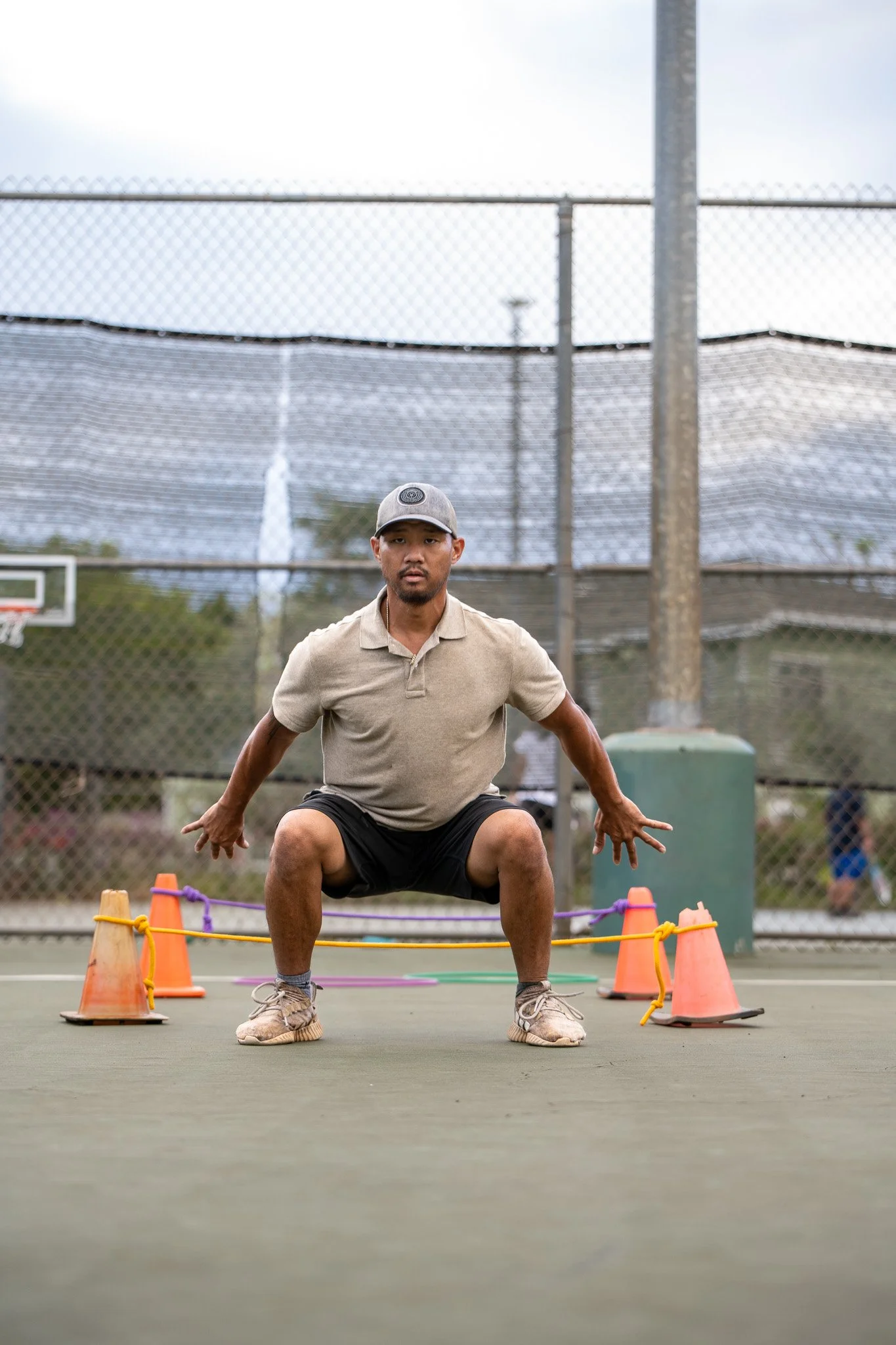 Man doing agility drills on an outdoor tennis court, squatting between orange cones, wearing a cap, beige polo, and black shorts.