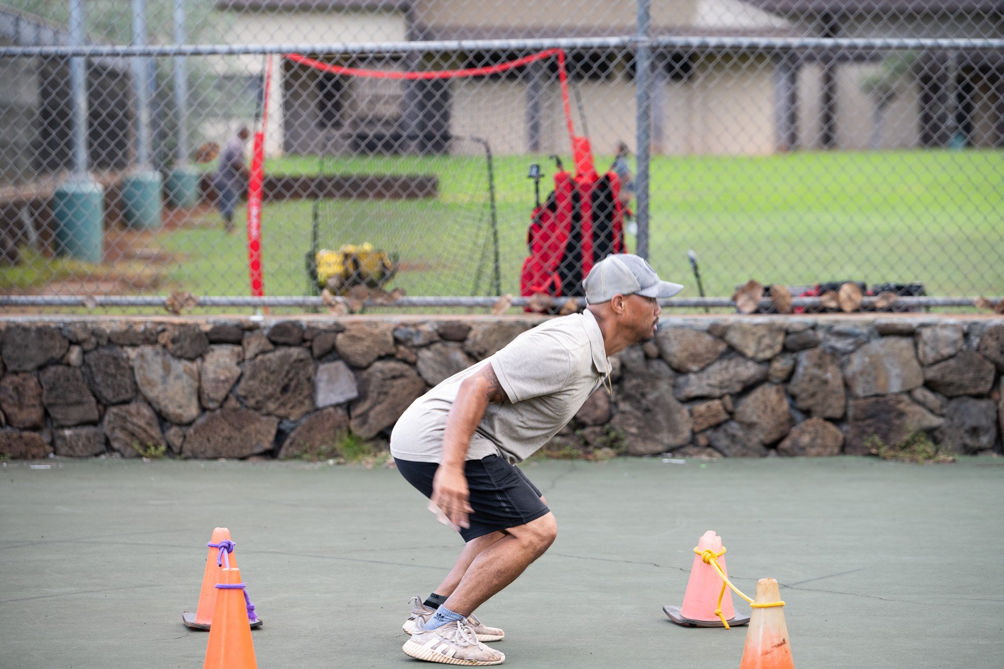 A man in a gray cap, beige t-shirt, black shorts, and sneakers is crouching on a green outdoor surface between two orange traffic cones tied together with purple and yellow ropes, indicating an agility or training drill.