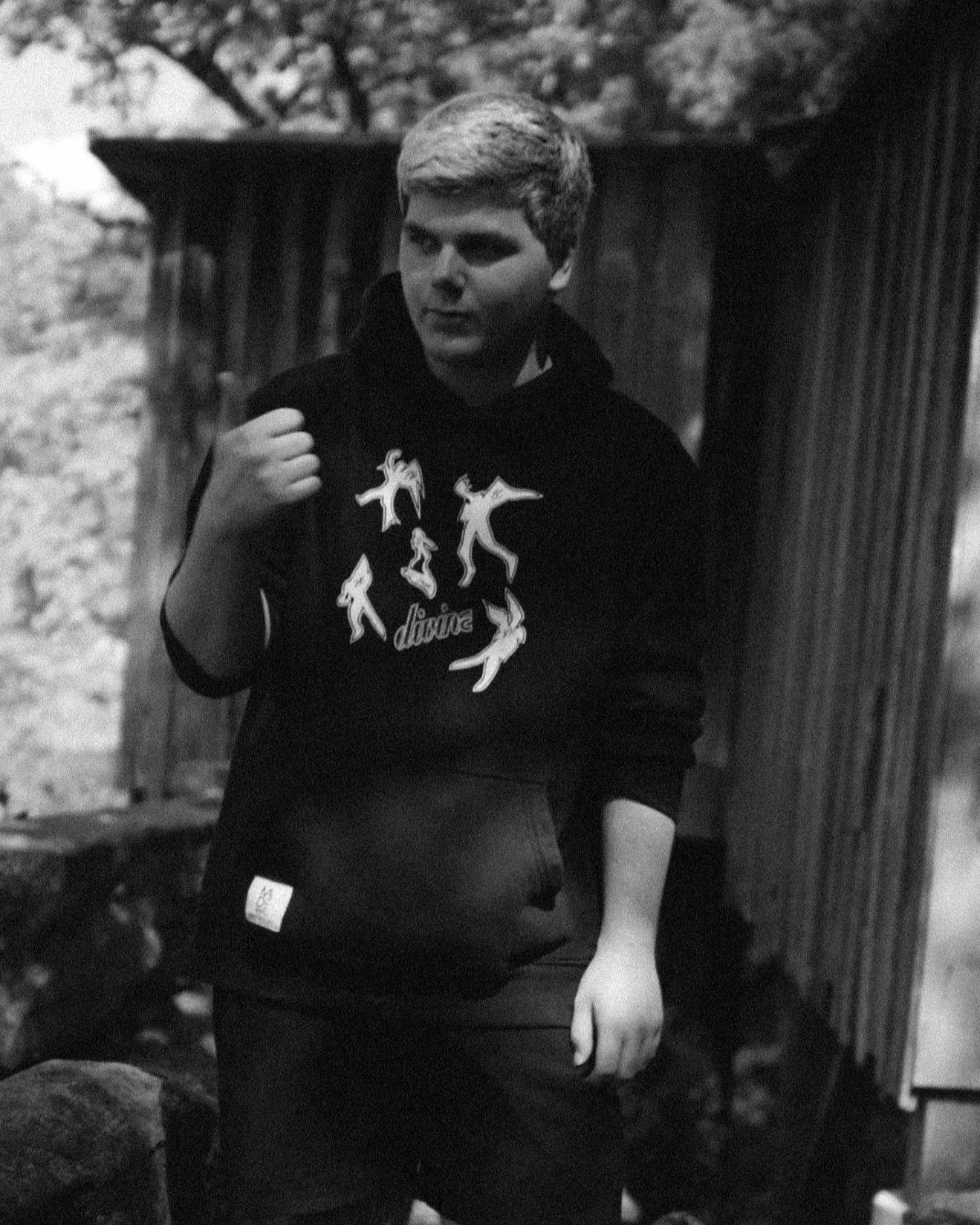 A young man with short blonde hair wearing a black hoodie with a graphic of dancing figures and the word 'dive' on the front, standing outdoors near a wooden fence, carrying a backpack.