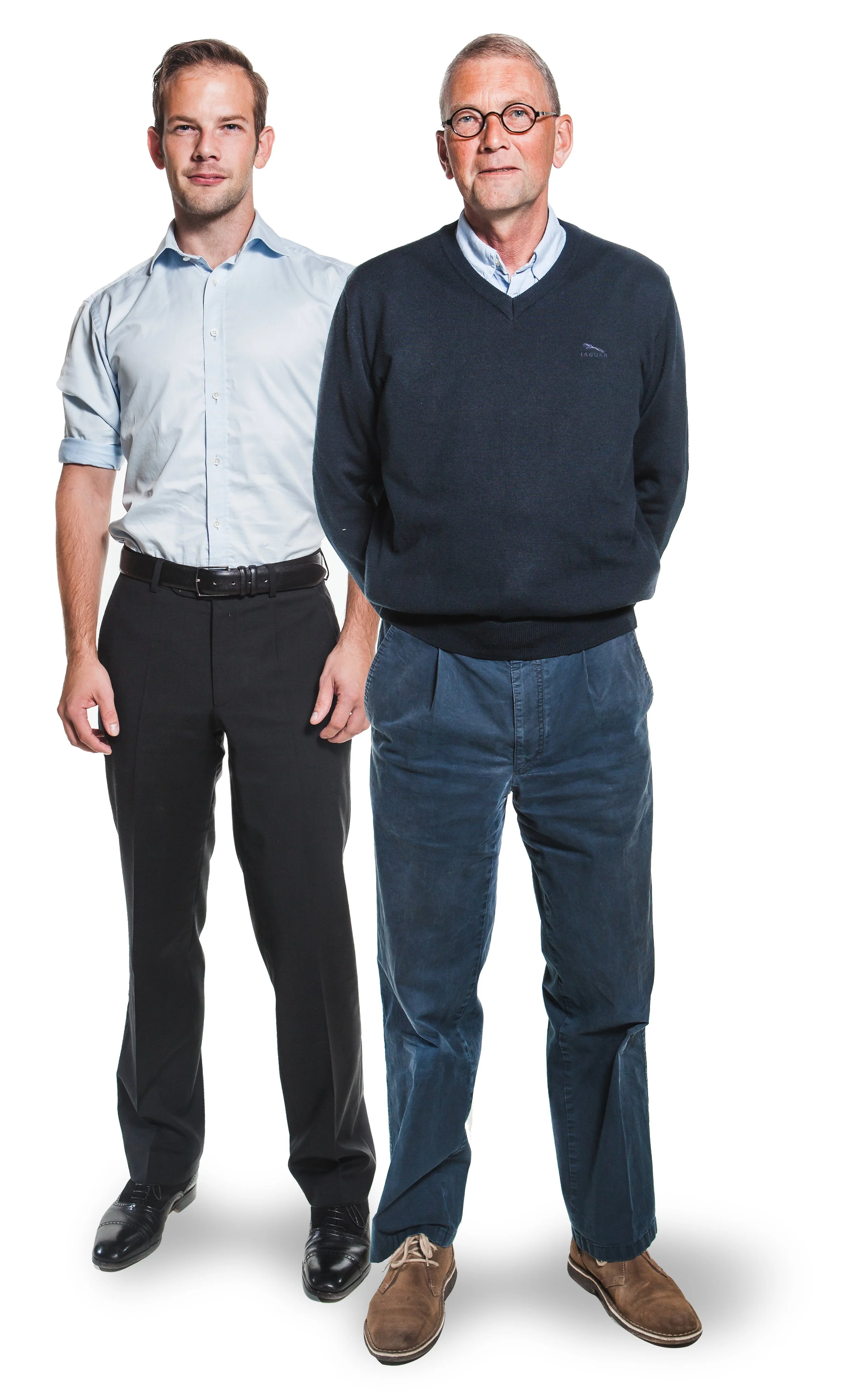 Two men standing against a plain white background, both wearing casual business attire. The younger man on the left wears a light blue shirt and dark pants, and the older man on the right wears a navy sweater, blue pants, and glasses.