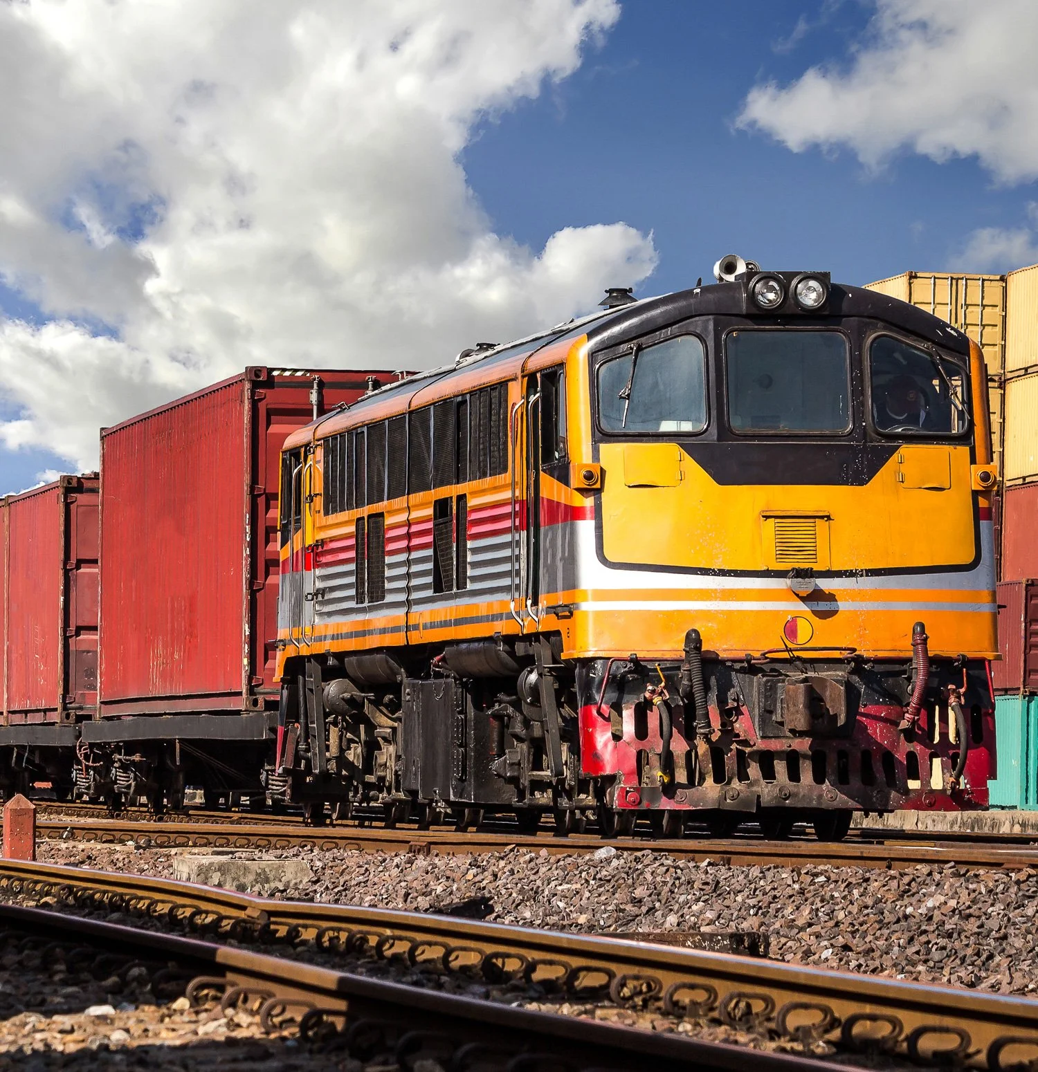 A yellow and black locomotive train pulling red freight containers, traveling on railway tracks under a partly cloudy sky.