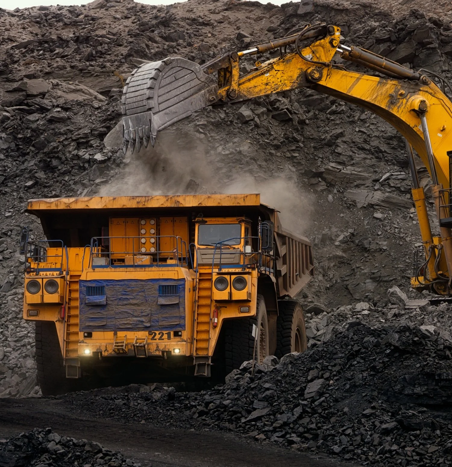 Heavy yellow dump truck being loaded with dirt or rocks by a large excavator on a rocky quarry or mining site.