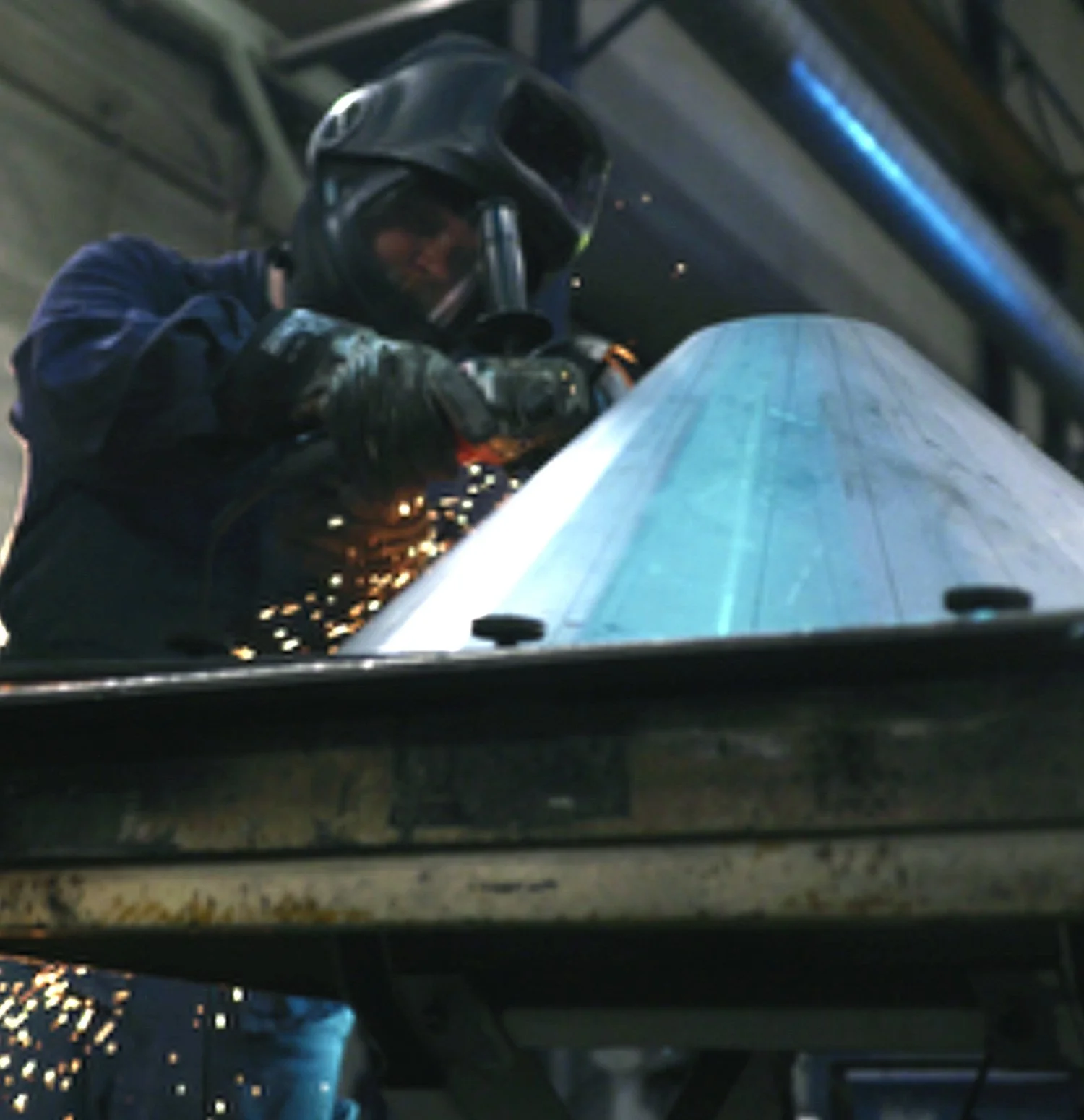 A worker wearing protective gear, including a welding helmet, is welding a large metal cone, with sparks flying from the welding process in an industrial setting.