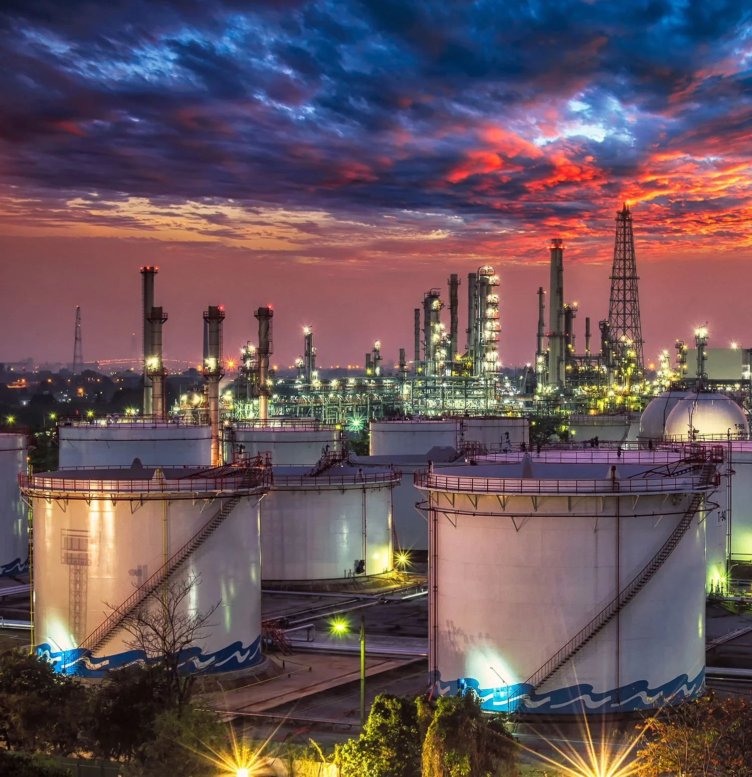 An industrial oil refinery at sunset with storage tanks in the foreground and distillation columns and towers in the background, against a colorful sky.