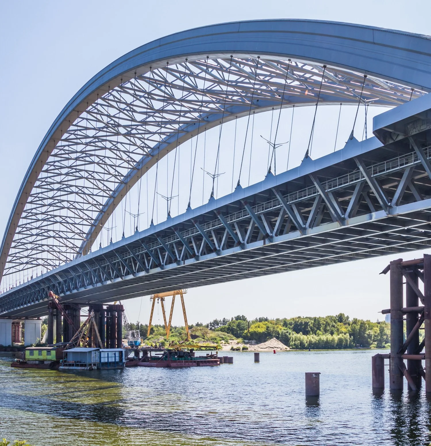 Construction of a modern steel arch bridge over a river on a sunny day with a clear blue sky and green trees in the background.