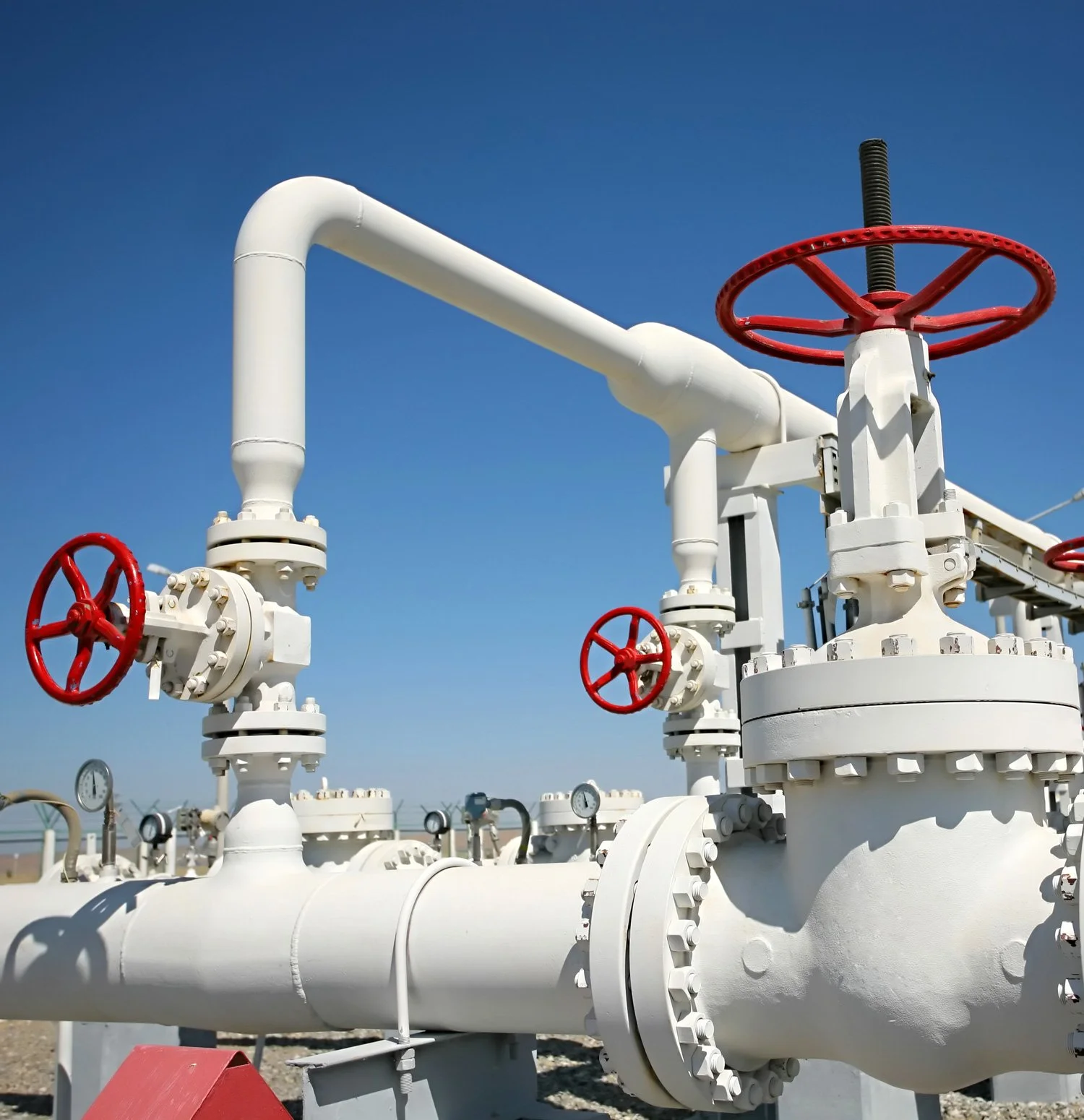 Industrial pipeline system with red valve wheels and pressure gauges against a clear blue sky.