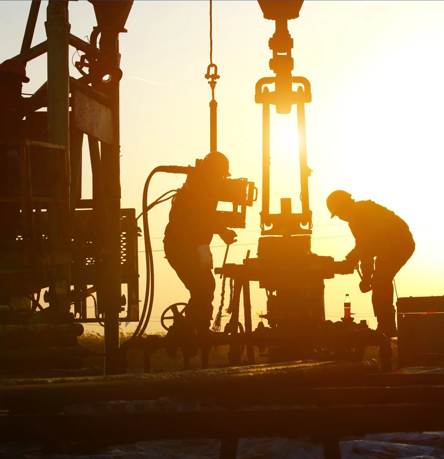 Silhouettes of two oilfield workers working on drilling equipment at sunset.