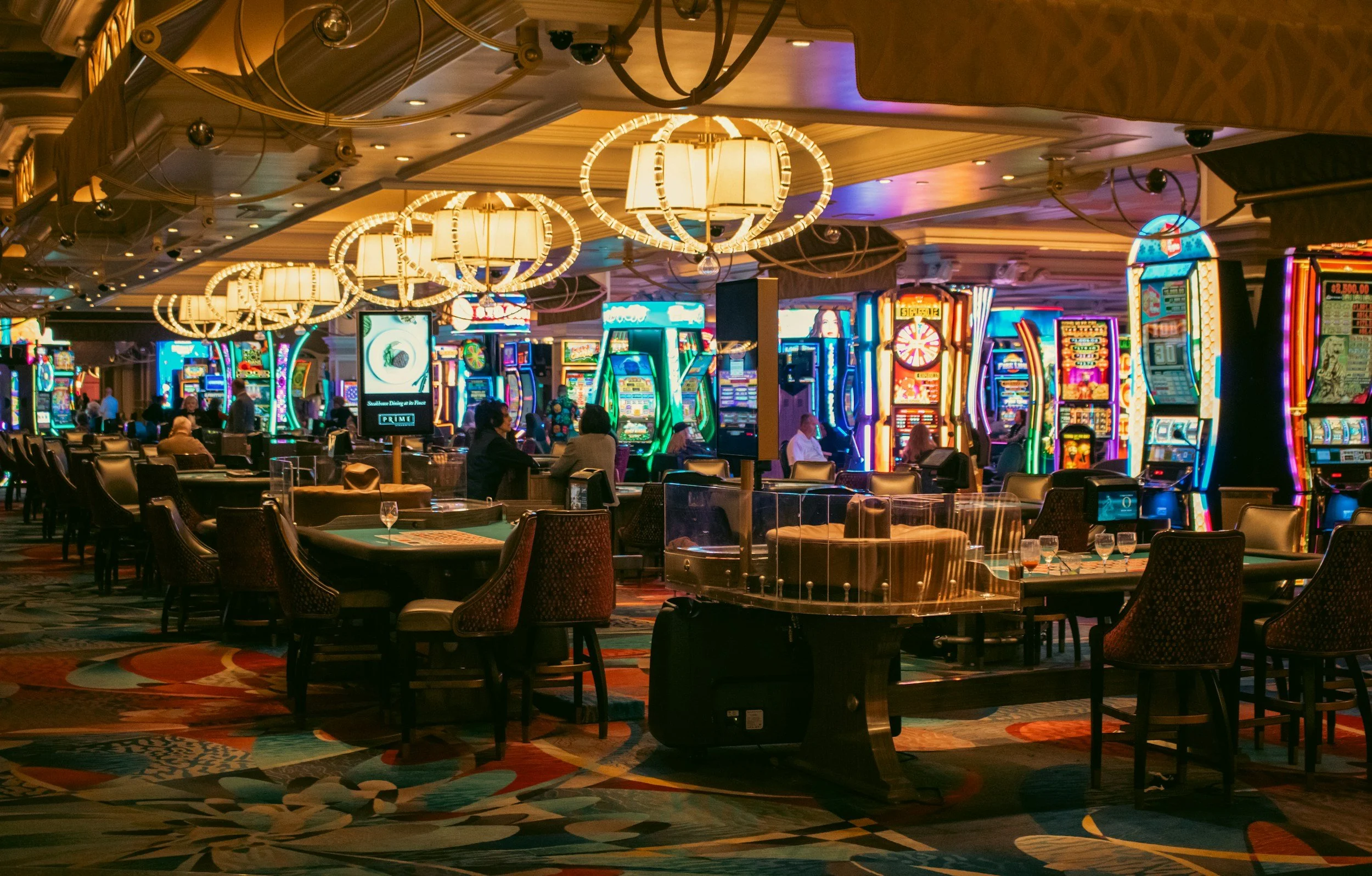 Interior of a casino with slot machines, gaming tables, and people playing, illuminated by colorful neon lights and chandeliers.