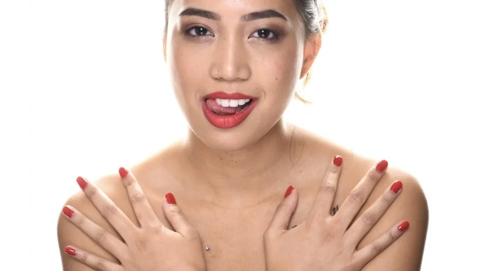 A young woman with red lipstick and nails, smiling with her tongue slightly out, against a white background.