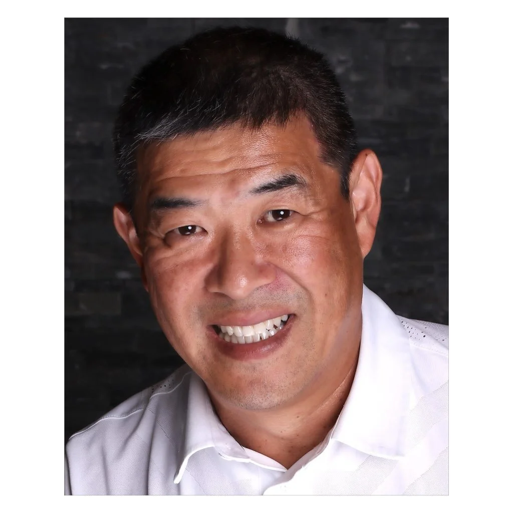 A smiling middle-aged Asian man with short black hair and a white collared shirt, posing against a dark, textured background.
