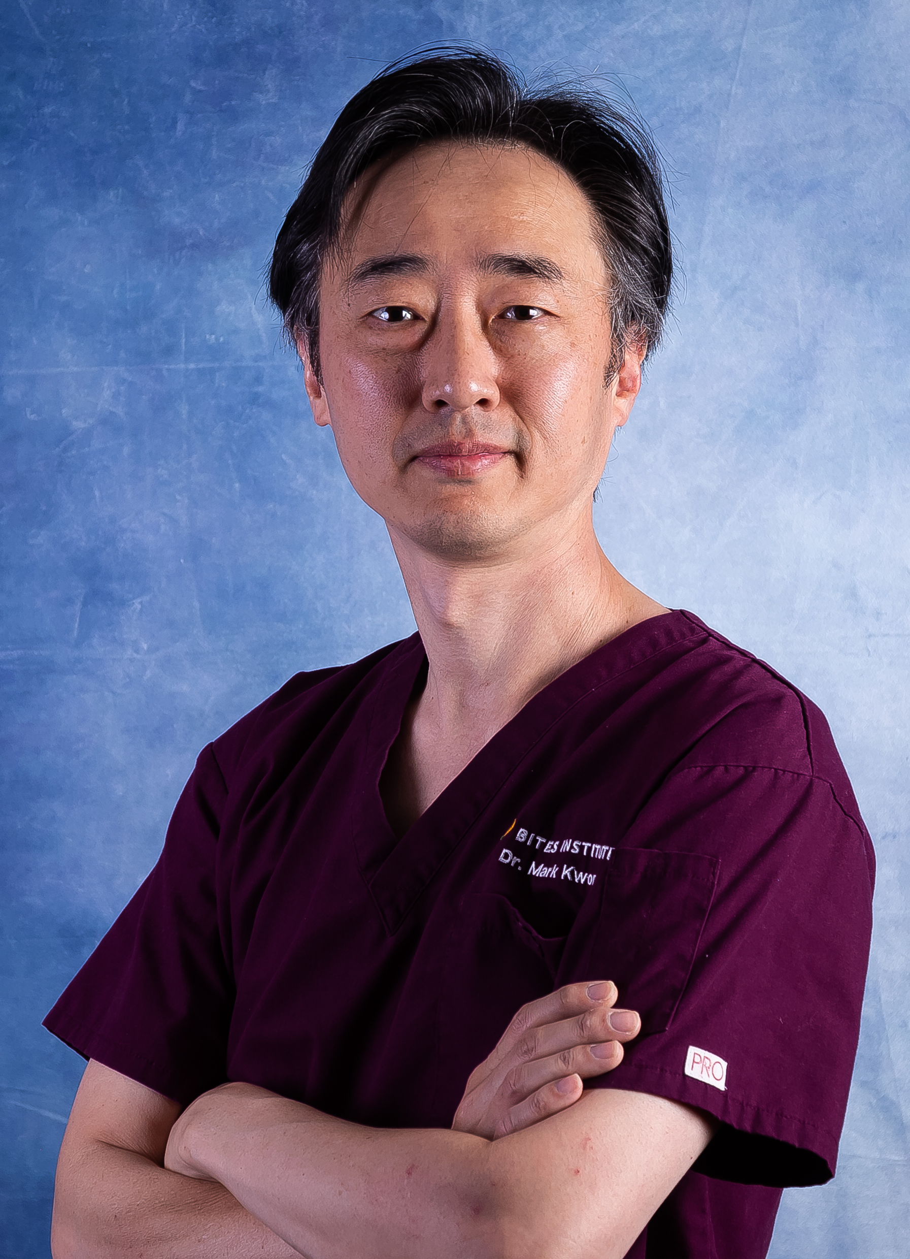 A man in a burgundy medical uniform with arms crossed, standing against a blue background.