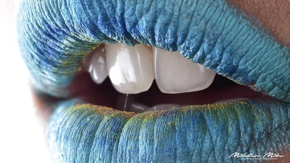 Close-up of lips painted with blue, green, and yellow lipstick, partially open to reveal white teeth.