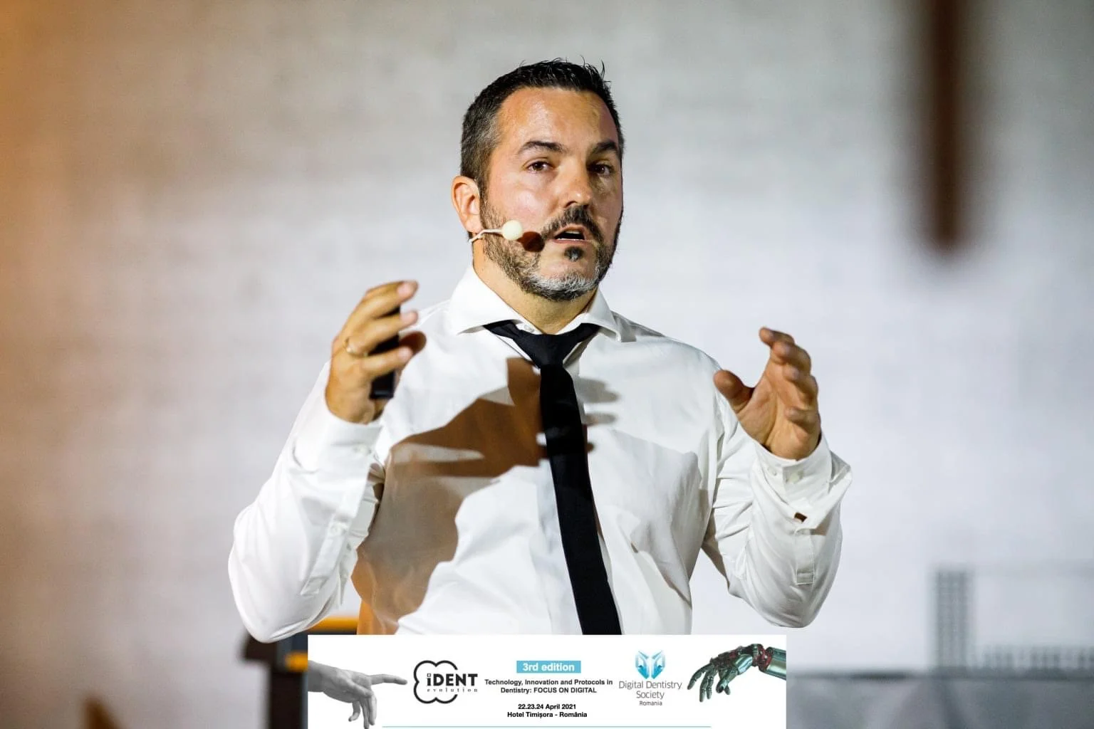 A man with dark hair, a beard, and mustache, wearing a white shirt and black tie, speaking into a headset microphone during a presentation.