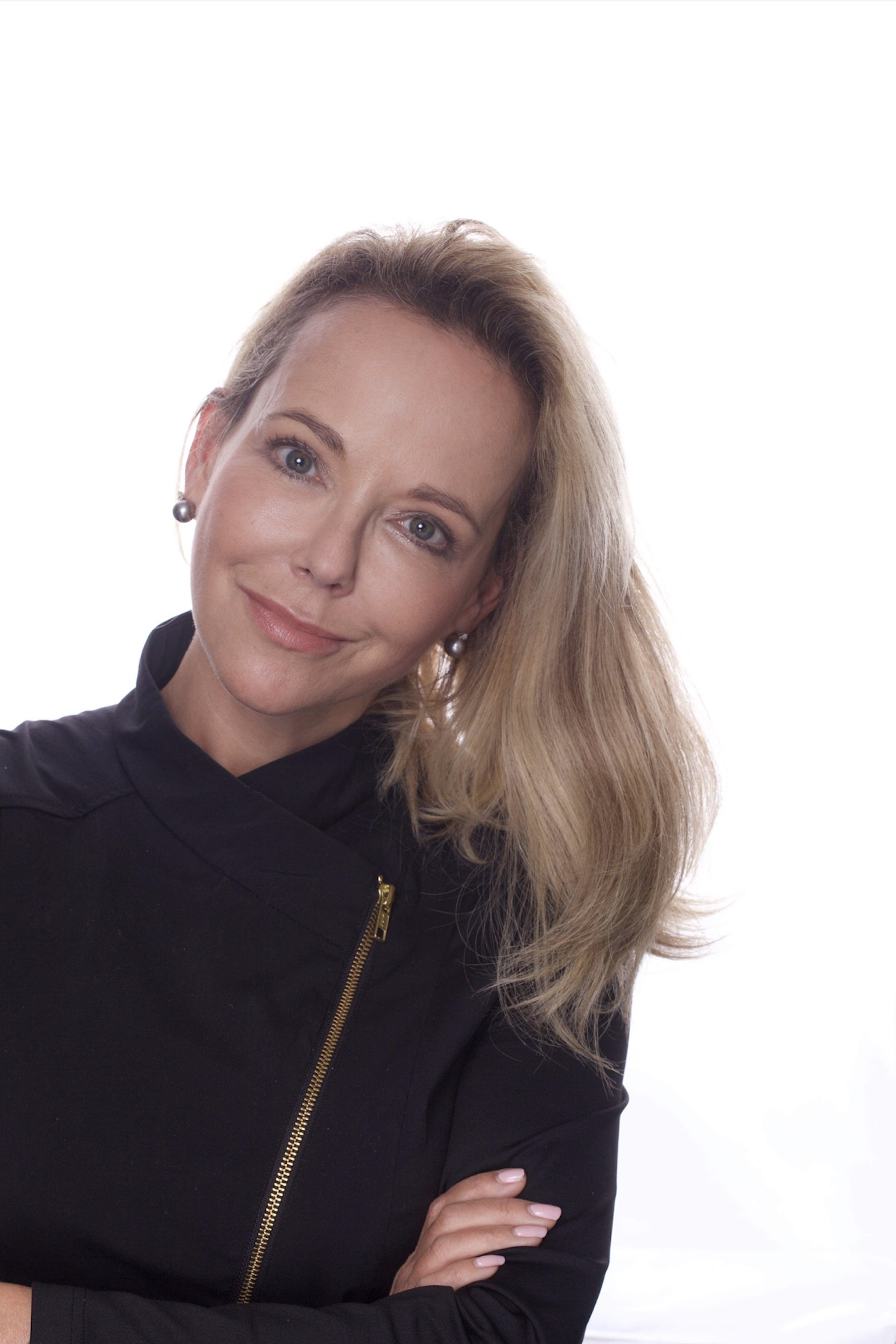 Close-up of a woman with blonde hair and blue eyes, wearing a black jacket and standing with her arms crossed, smiling softly, against a white background.