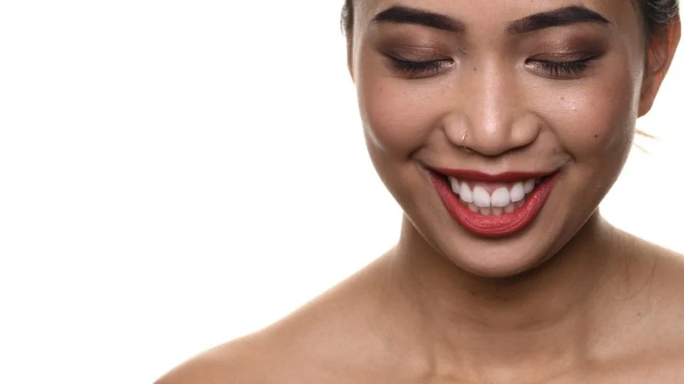 Close-up of a smiling woman with closed eyes, red lipstick, and a nose ring, against a white background.