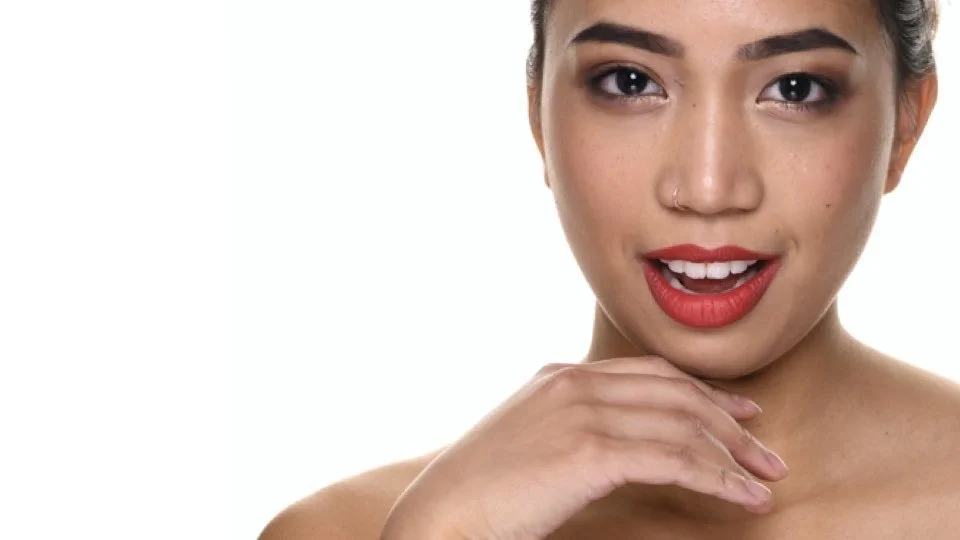 Close-up of a woman with flawless skin, red lipstick, and a small nose piercing, posing against a white background.