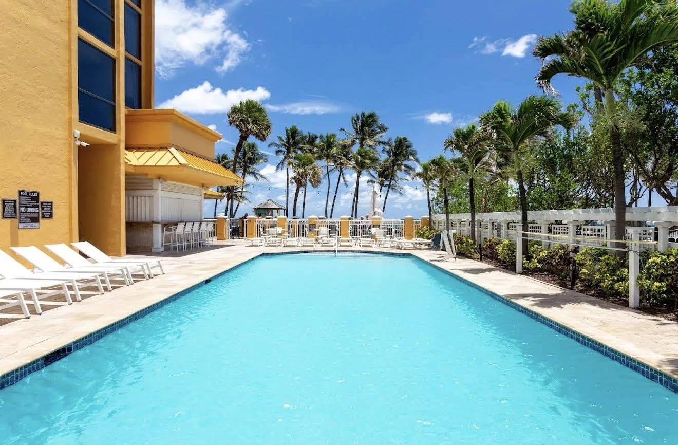 A backyard swimming pool area with lounge chairs, surrounded by palm trees and a bright blue sky.