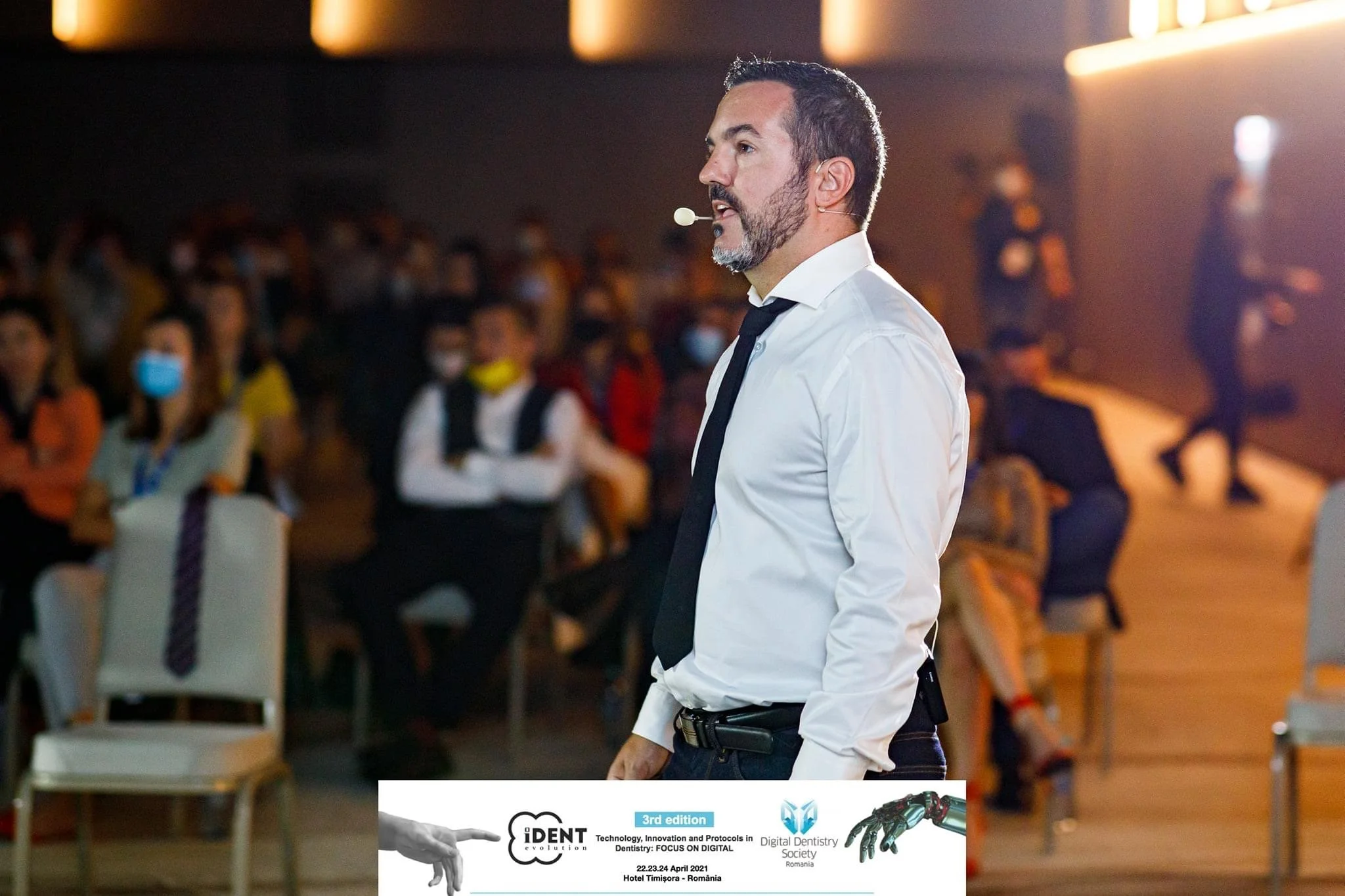 A man with dark hair and beard, wearing a white shirt and black tie, speaking at a conference with a microphone headset in front of an audience, with a conference banner at the bottom.