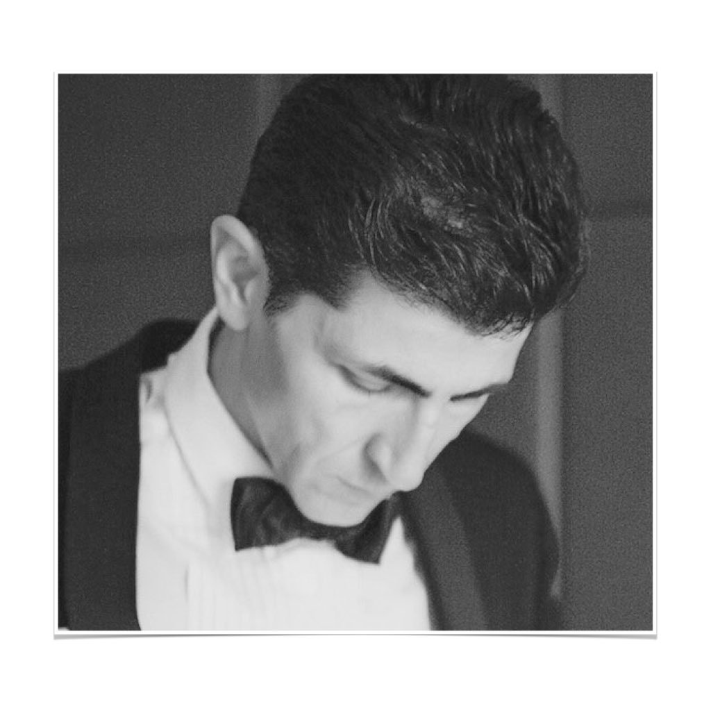 Black and white photo of a man dressed in formal attire, with a tuxedo and bow tie, looking downward with a serious expression.