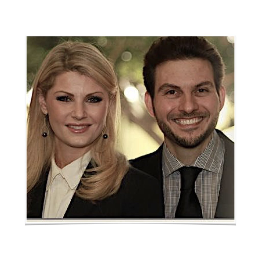 A smiling woman with blonde hair and earrings standing next to a smiling man with dark hair and a beard, both dressed in business attire, outdoors with blurred trees in the background.